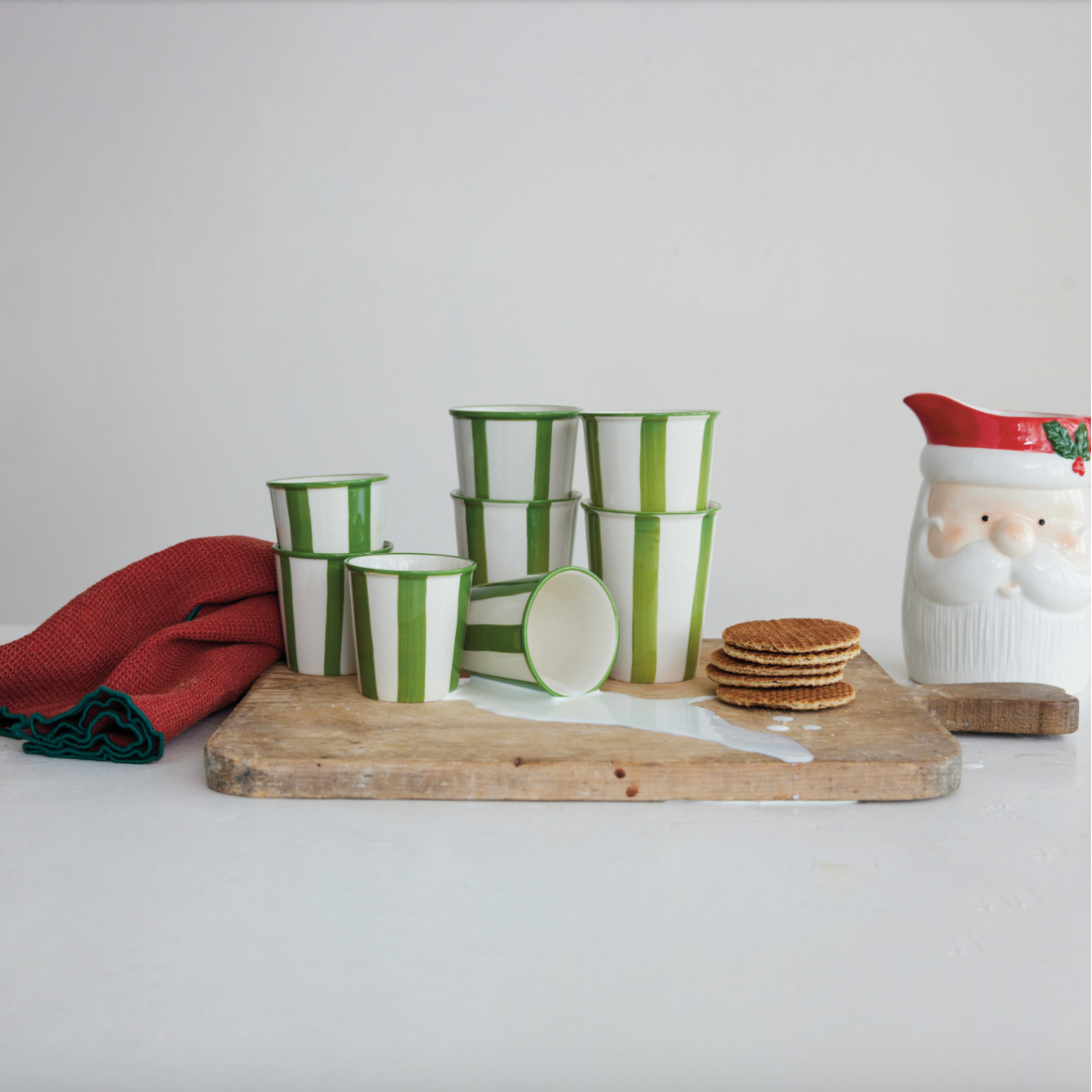A set of green and white striped cups arranged on a wooden board, alongside a red tea towel, stacked round cookies, and a white Santa-themed pitcher on a plain light background.