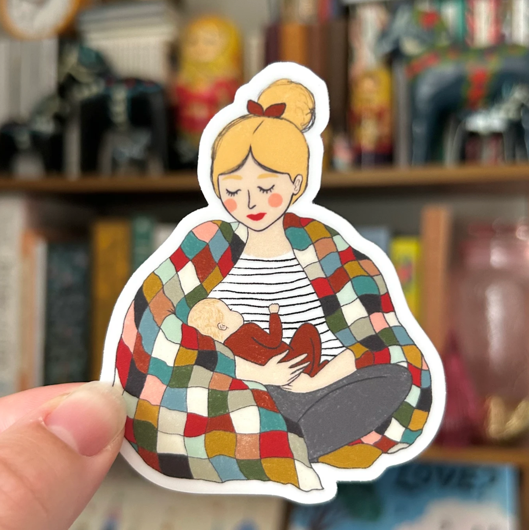 A mother with blonde hair and fair skin wearing a black and white striped top is holding a fair skinned baby wearing a deep red onesie.