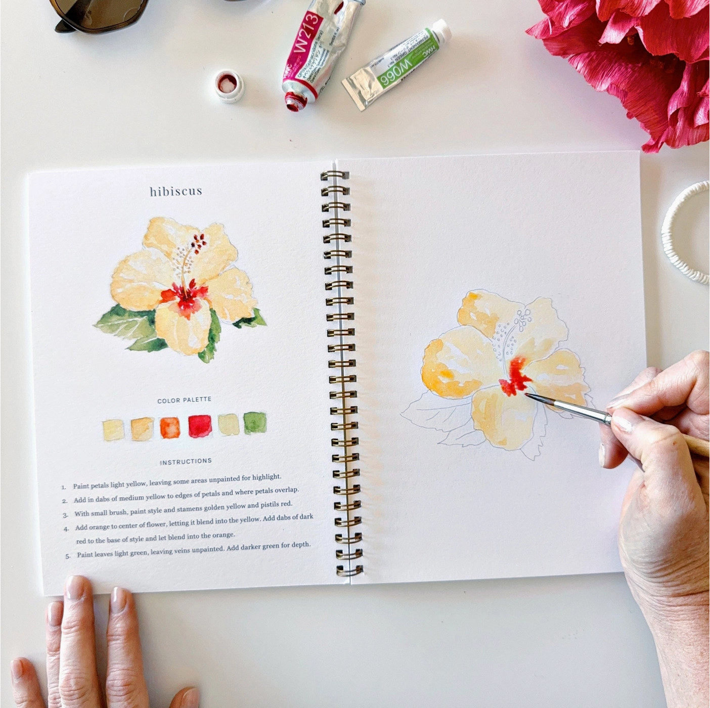 The watercolor book open with a scene of a hibiscus flower and instructions to paint it.