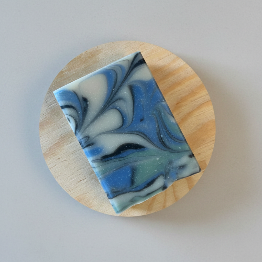 A rectangular bar of marbled soap in blue, white, and black swirls, displayed on a round wooden coaster against a blurred green background with raindrops.