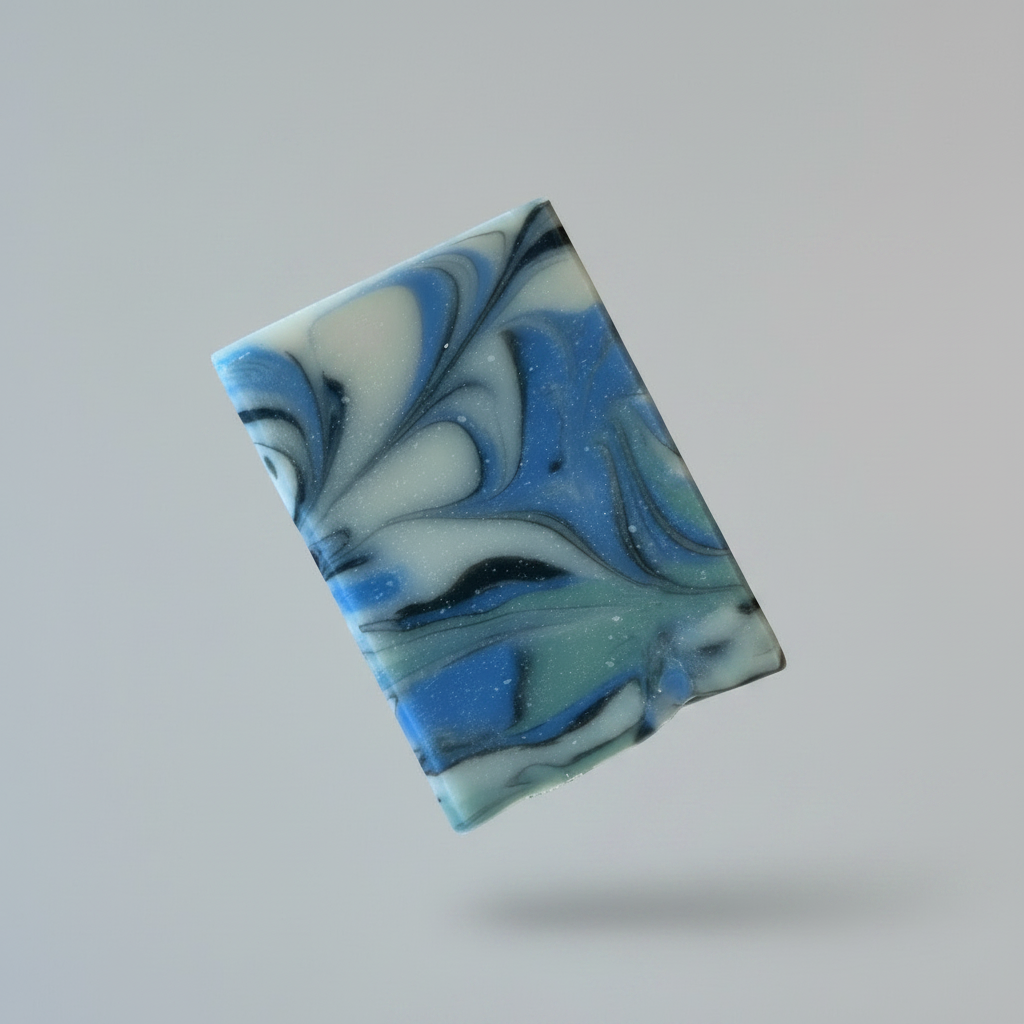 A rectangular bar of marbled soap in blue, white, and black swirls, displayed on a round wooden coaster against a blurred green background with raindrops.