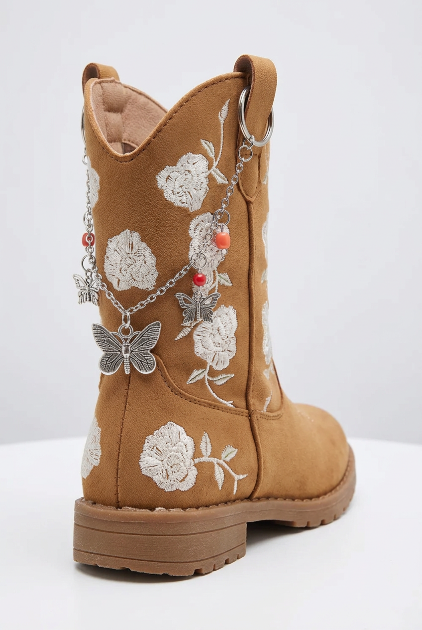 Tan toddler cowboy boot with cream floral embroidery, adorned with silver-tone chain charms featuring a butterfly pendant, a floral charm, and small red bead accents.