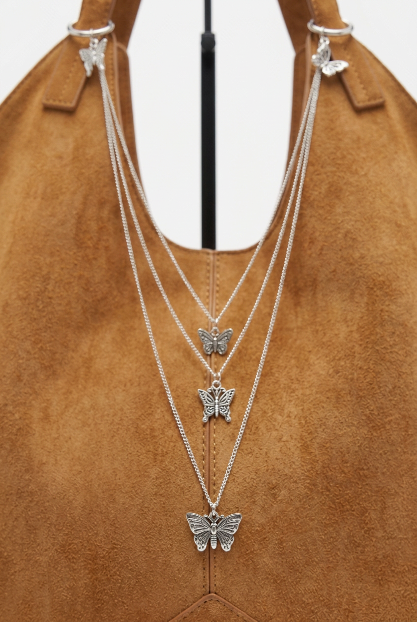 Tan suede handbag adorned with layered silver-tone chains and multiple intricate butterfly charms, creating a delicate, whimsical accessory.