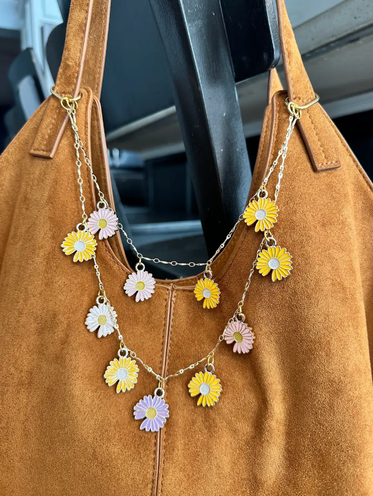 Tan suede handbag adorned with layered gold-tone chains and colorful daisy charms in yellow, pink, white, and lavender, featuring bright floral pendants with contrasting centers.