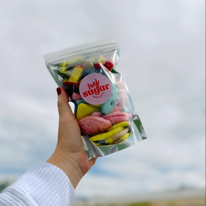 A hand holding a clear bag of colorful assorted candies with a round pink label that reads “hej sugar” against a cloudy sky background