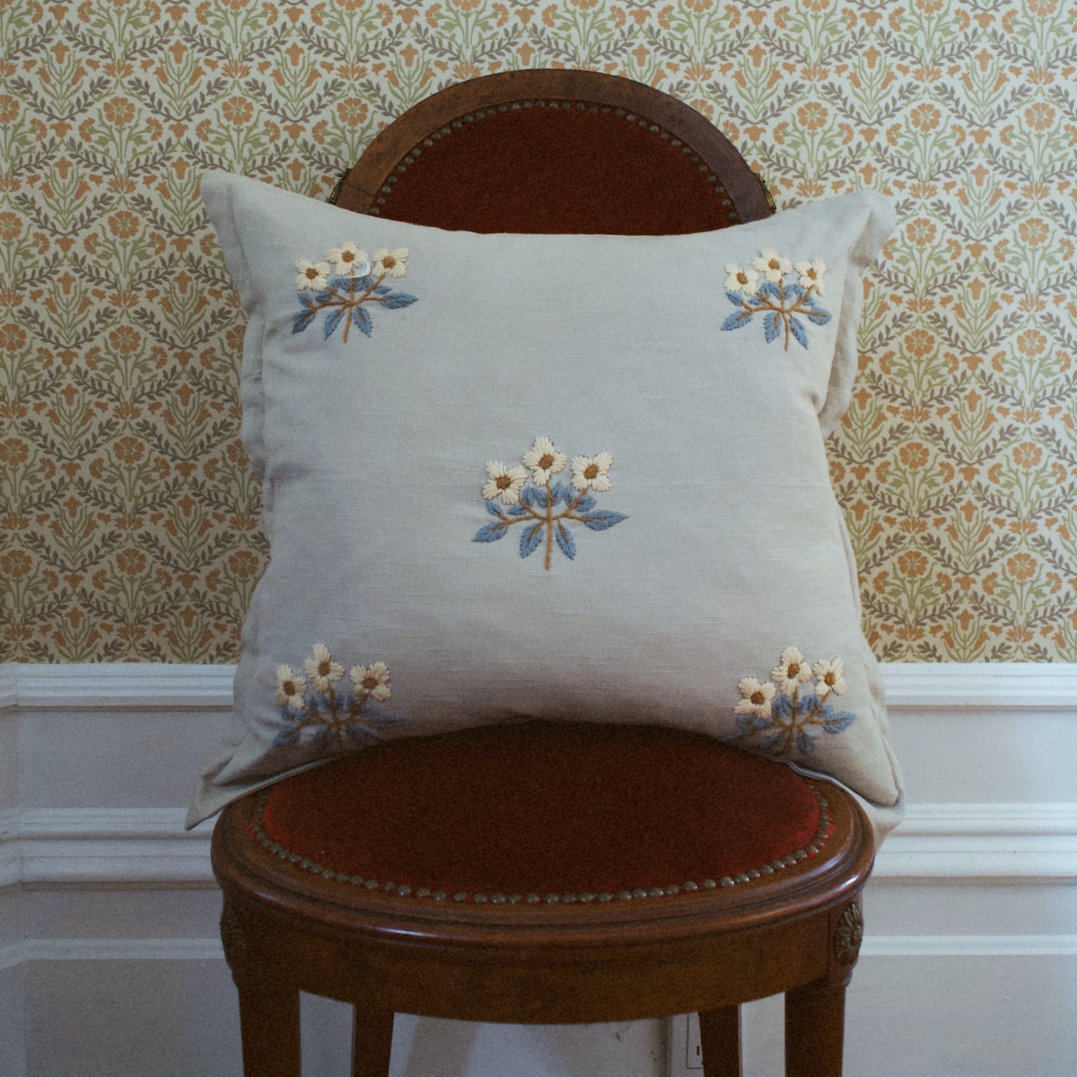 A beige pillow with a floral pattern, featuring five embroidered flowers, placed on a chair with a patterned wall in the background.