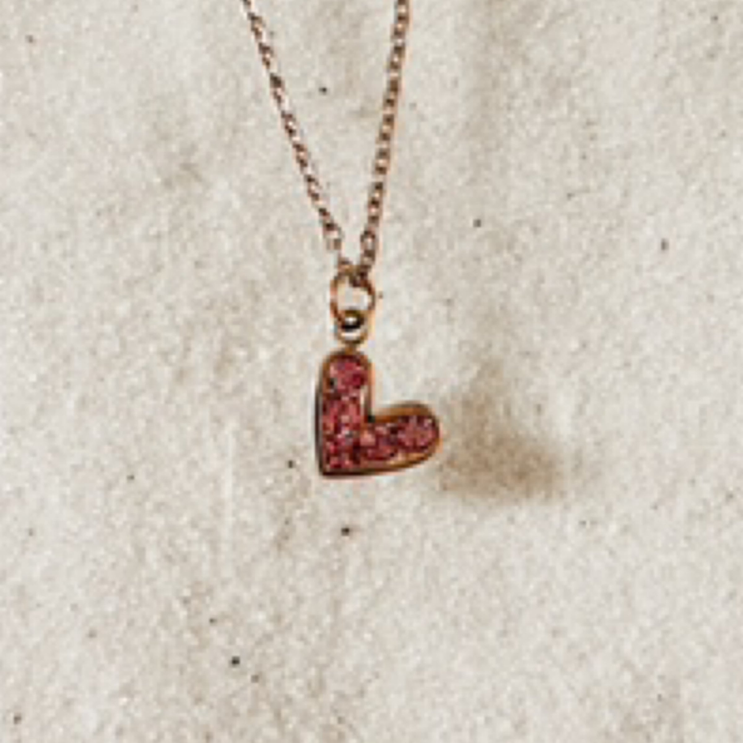 Delicate gold chain with a gold rimmed pink gem stone heart charm hanging on a tilt. Pictured against white sand.