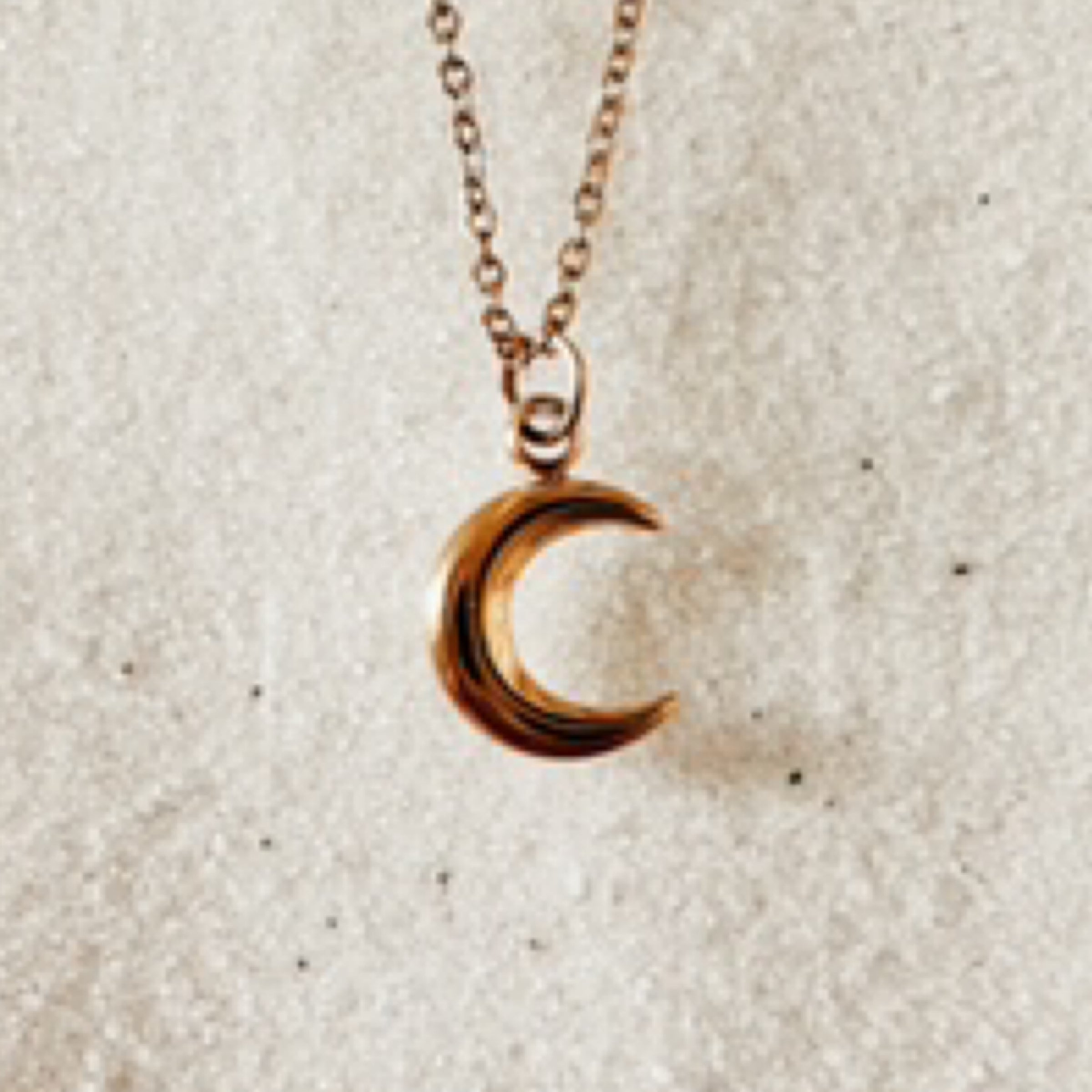 Delicate gold chain with a small gold crescent moon charm. Pictured against white sand.