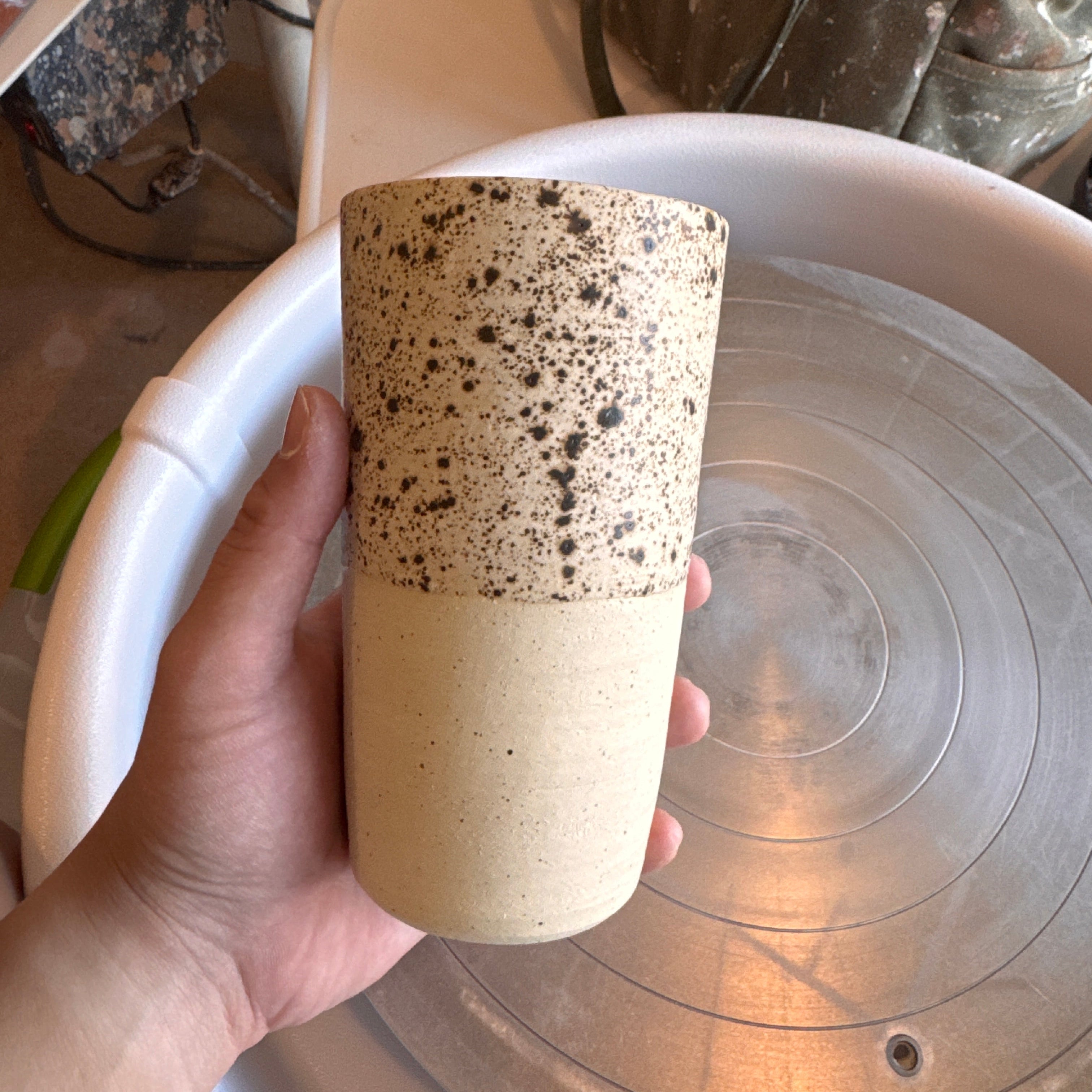 A hand holds a handmade ceramic tumbler with a speckled black-on-cream glaze covering the upper half and a smooth plain cream lower half, displayed in front of a pottery wheel. The background shows a pottery studio workspace with clay tools, bats, and scattered equipment on the wheel.