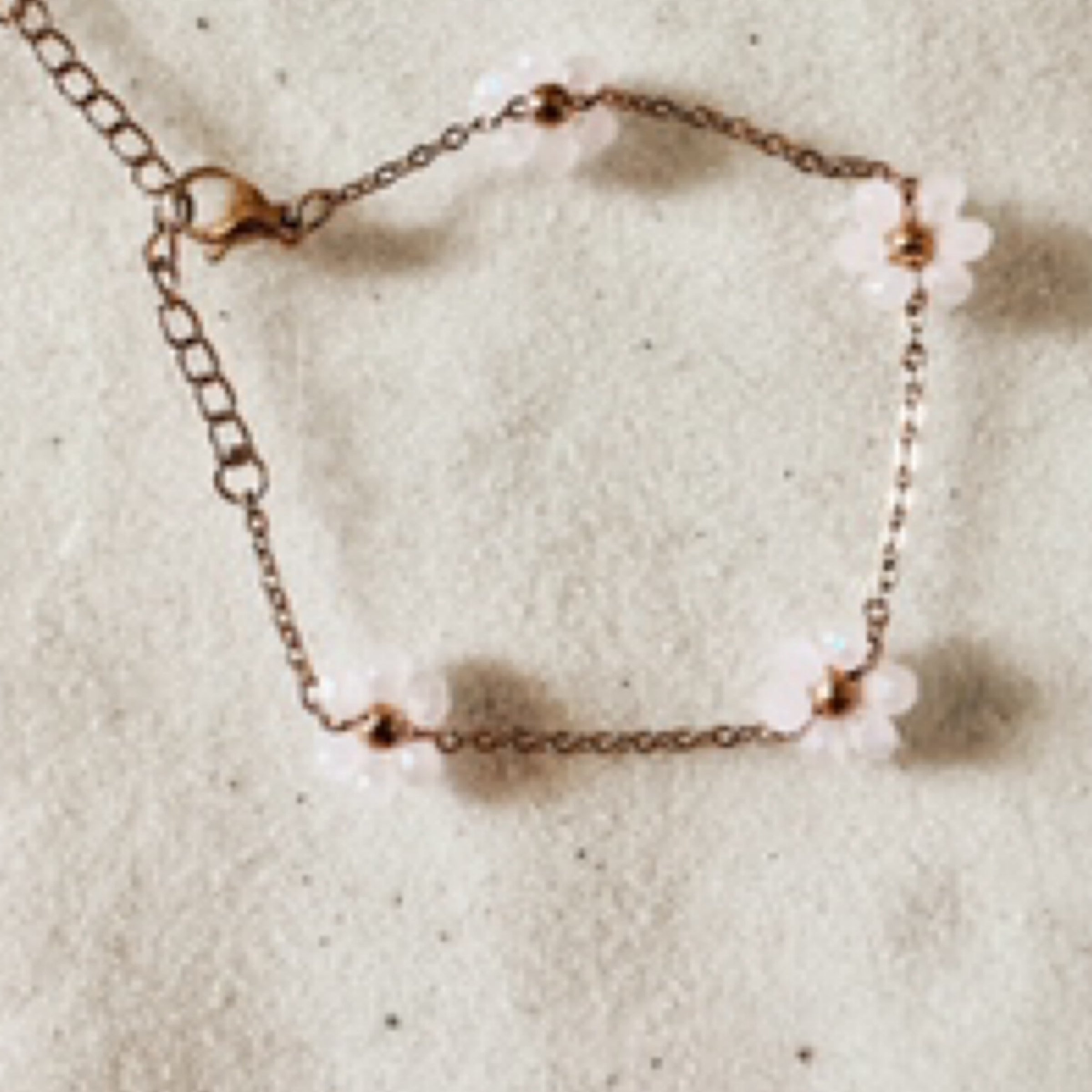 Delicate gold chain with light pink colored sparkly stones clustered together like daisy petals around a gold bead as the center of the daisy. (total of 4 daisies per bracelet). Pictured against white sand.