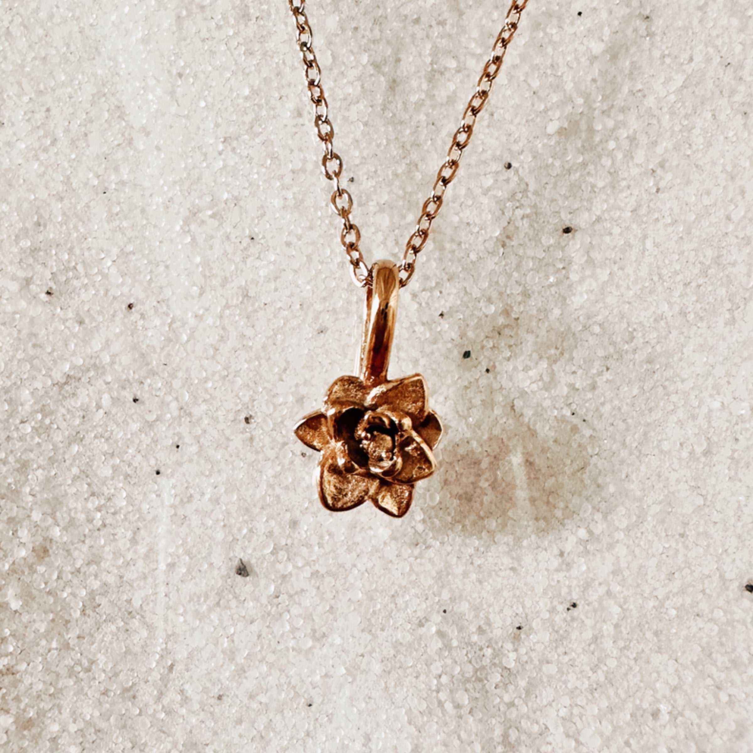 Delicate gold chain with a detailed gold daffodil charm. Pictured against white sand.