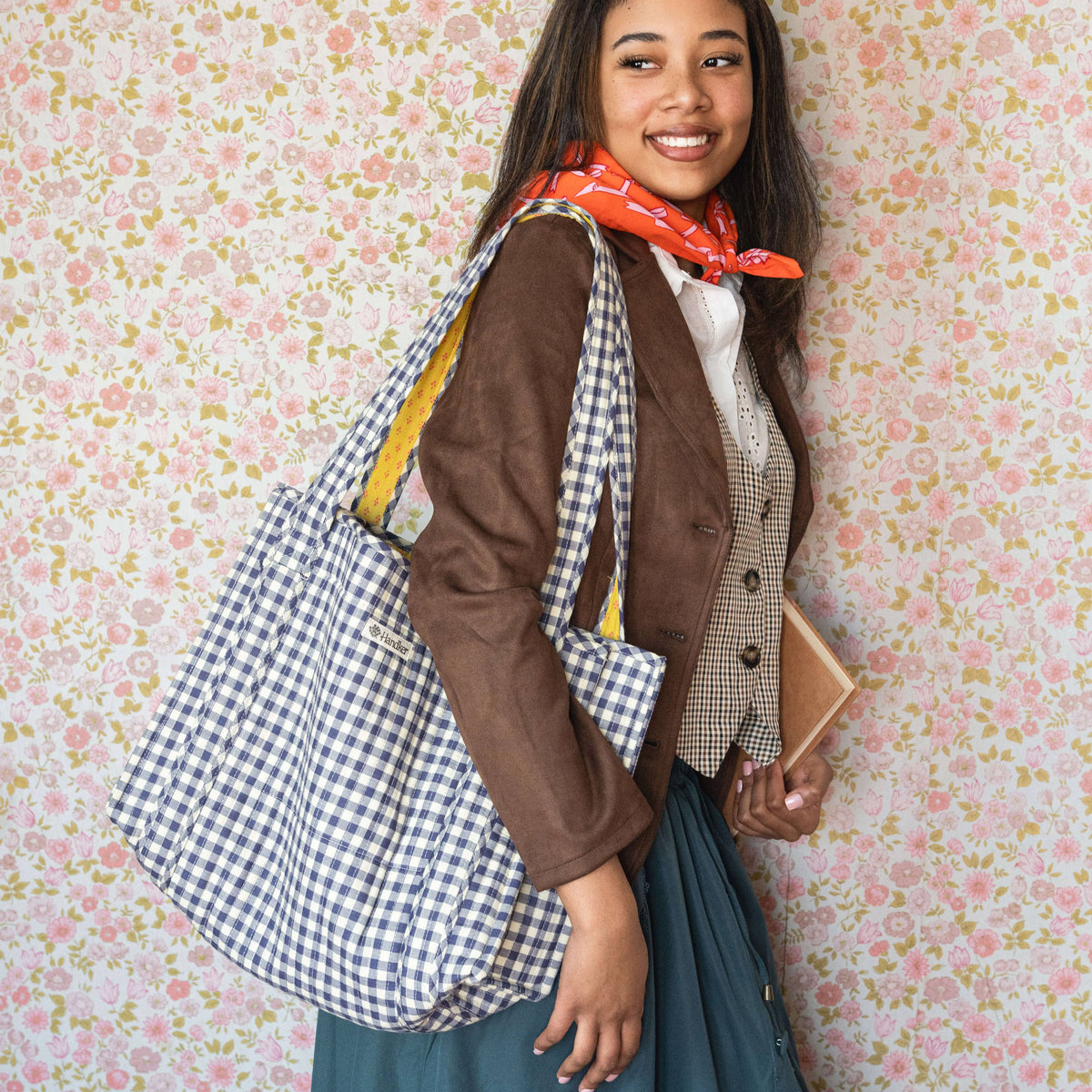 Woman holding a checkered bag and wearing a red scarf against a floral wallpaper background