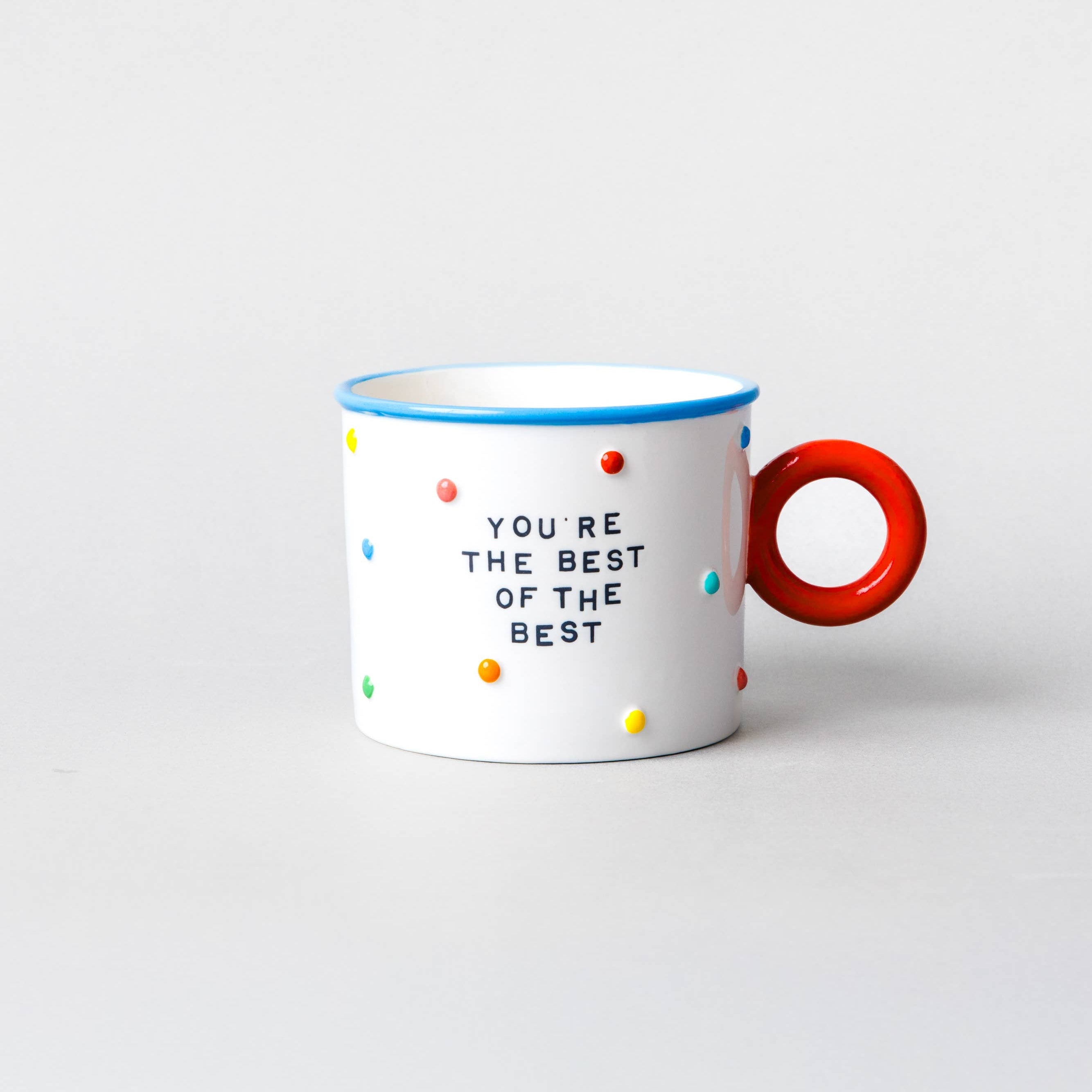 A white ceramic mug with a bright red handle, decorated with colorful polka dots and the text 'YOU'RE THE BEST OF THE BEST' in black. The mug has a blue rim and is set against a plain light gray background.