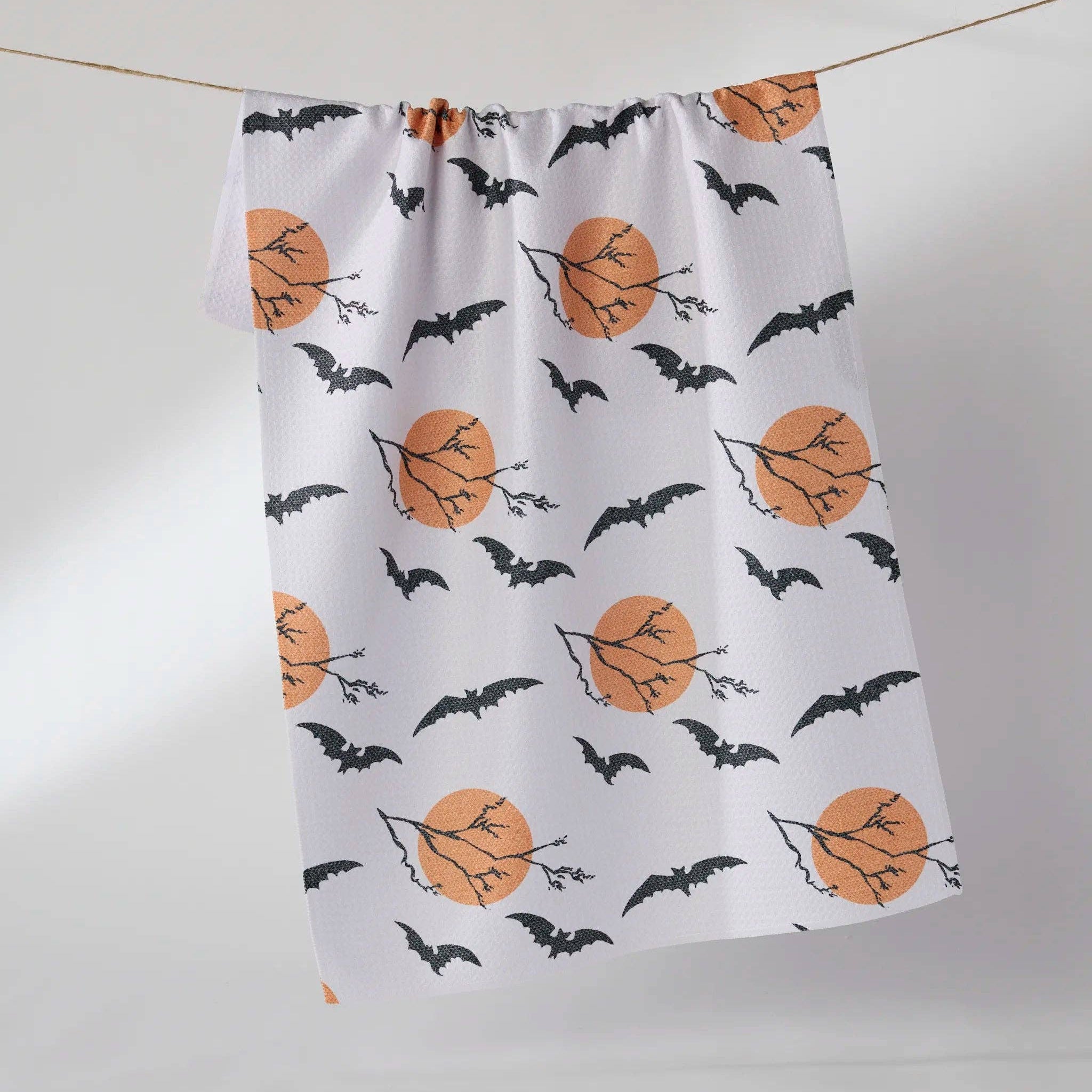 A white kitchen towel with a Halloween pattern of black bats, bare tree branches, and orange full moons, hanging from a string against a light gray background.