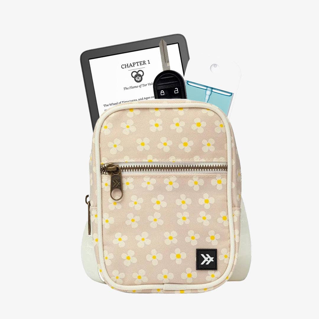 A small beige crossbody bag with a white and yellow daisy floral print, shown with its front zip pocket open and holding a tablet, keys, and a blue card. The bag has a black logo patch on the bottom right corner.