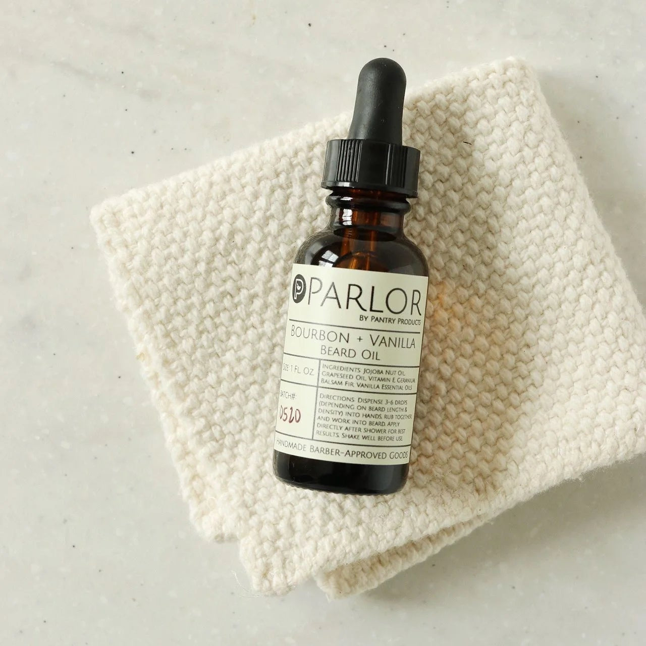 A brown glass dropper bottle labeled “Parlor by Pantry Products: Bourbon + Vanilla Beard Oil” rests on a folded cream-colored textured cloth. The label highlights natural ingredients like jojoba and grapeseed oil, and the overall presentation gives a clean, artisanal, and grooming-focused vibe.