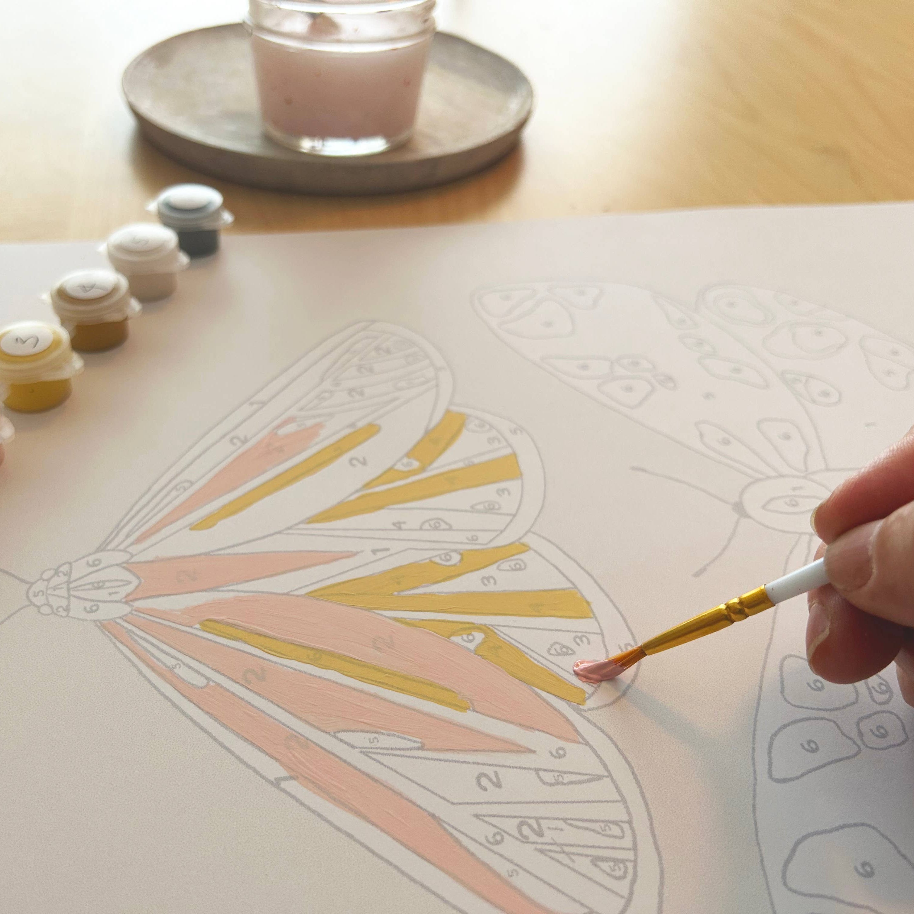 Person painting a butterfly design on paper with small paint pots and a candle in the background.
