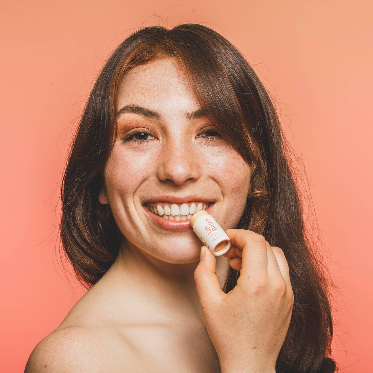 A smiling woman with dark brown hair applies a beige lip balm tube to her lower lip, set against a solid coral-pink background.