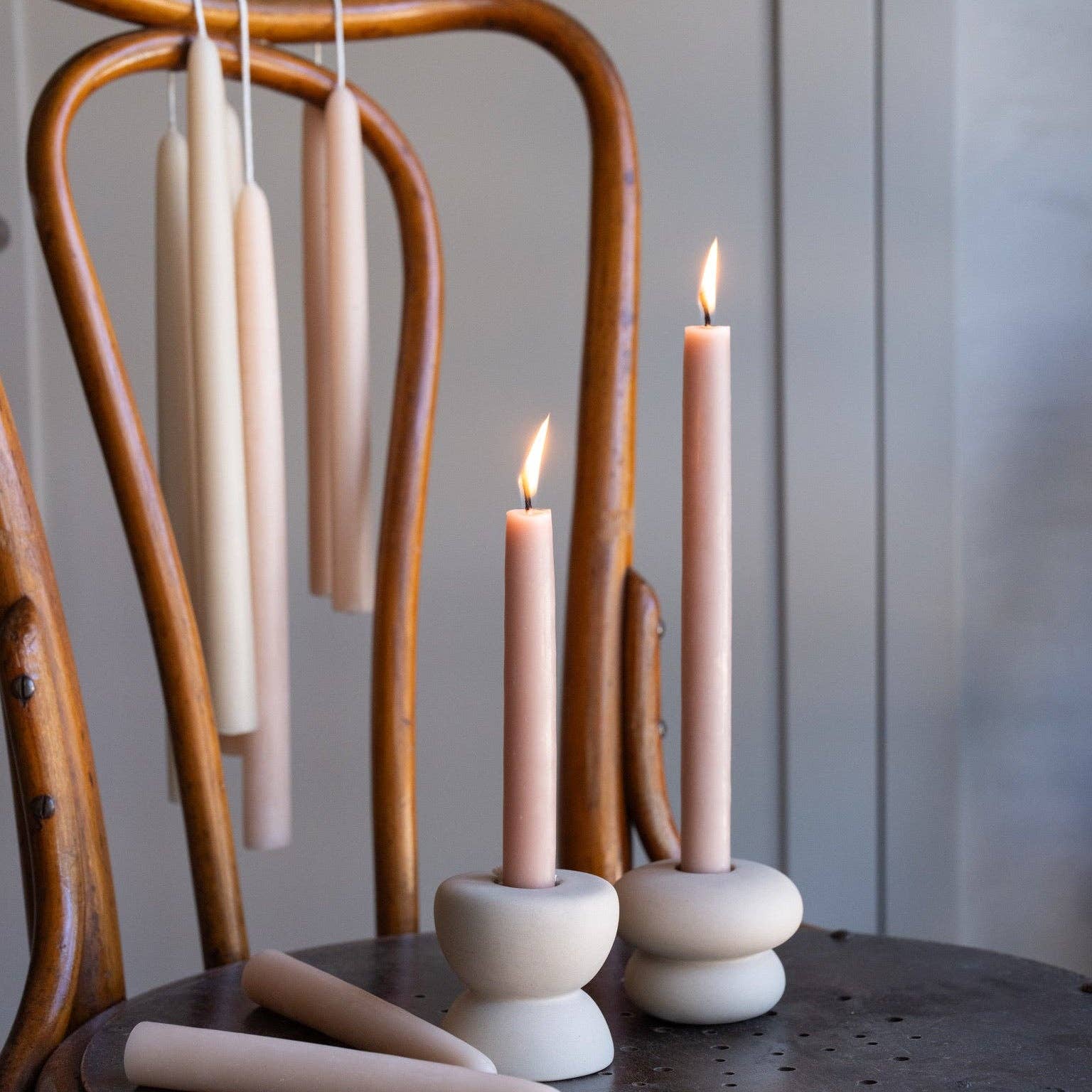 Two lit pale pink taper candles in round white ceramic holders on a dark wooden table, with additional unlit candles hanging on a curved wooden chair in the background.