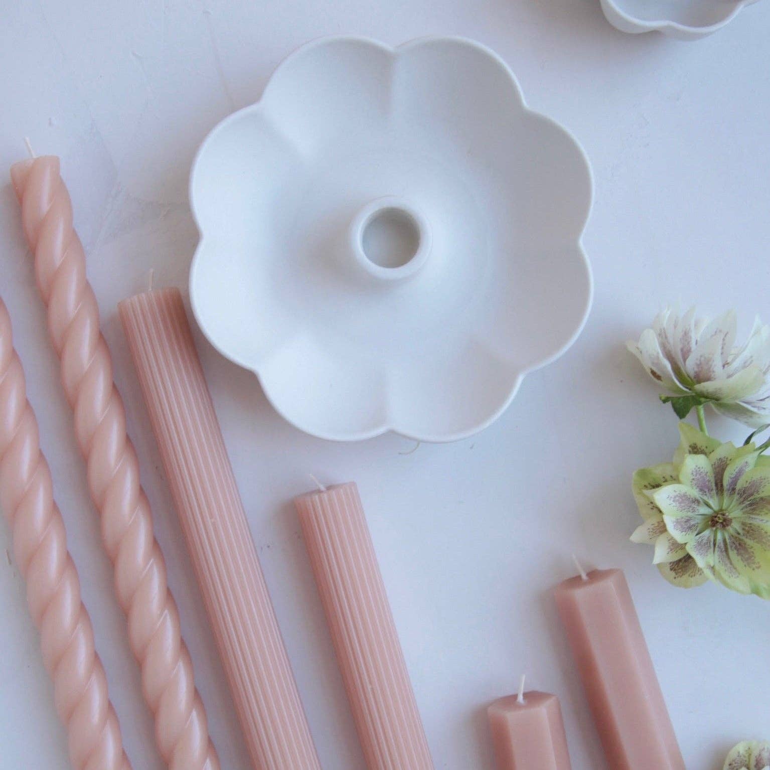 A white ceramic flower-shaped candle holder or mold displayed on a pale pink surface, surrounded by pastel pink twisted and ribbed candles and small decorative flowers.