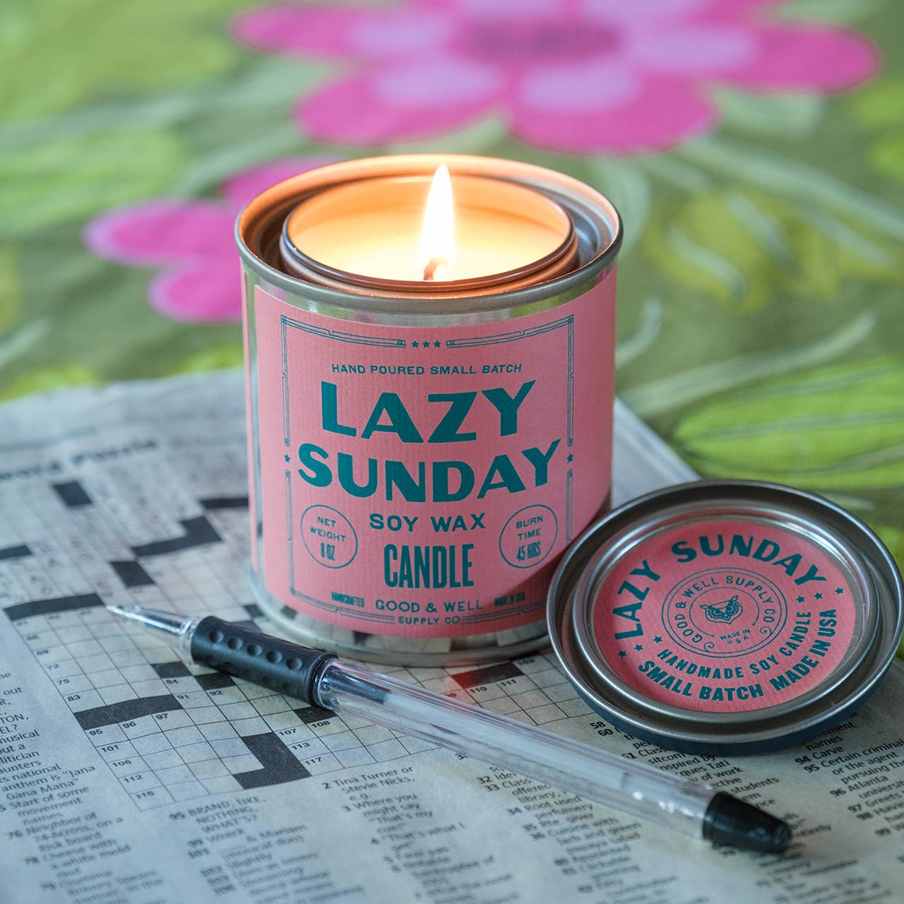 Candle in a pink container labeled 'Lazy Sunday' on a newspaper with flowers in the background