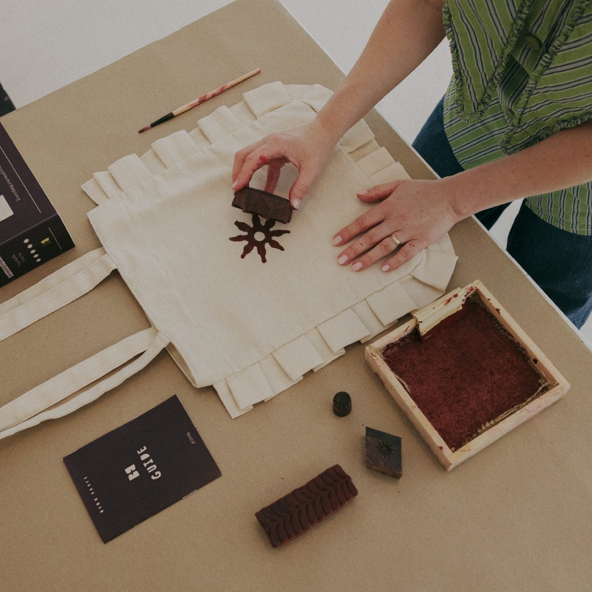 Person working with a stamp on fabric, surrounded by ink and a book on a beige surface.