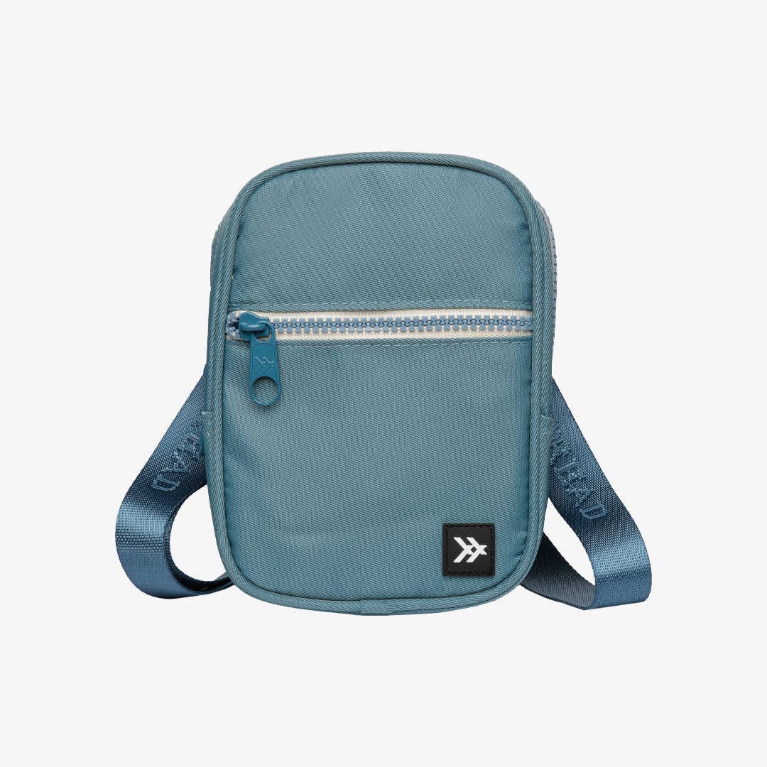 A small teal blue crossbody bag with a front zippered pocket, adjustable shoulder strap, and a small black brand patch on the lower right, shown against a light gray background.