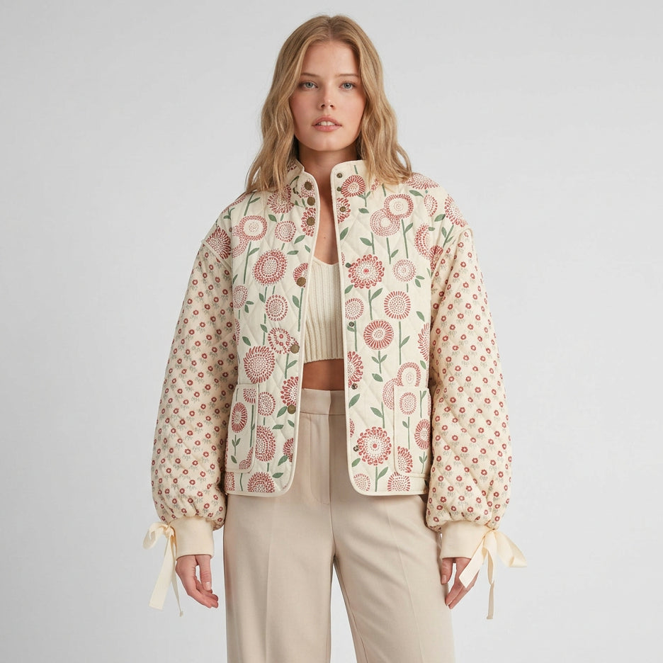 A woman modeling a cropped quilted jacket with a cream base, pink floral embroidery on the front, and pink dotted pattern on the sleeves, styled with brown drawstring pants against a warm beige background.