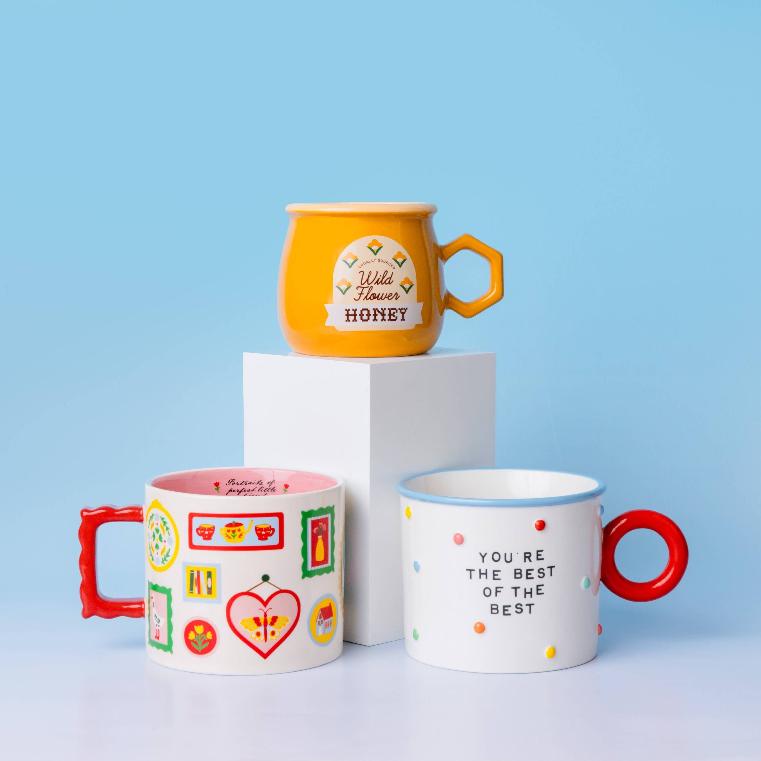 Three ceramic mugs displayed on a white pedestal and light surface against a soft blue gradient background. One yellow mug sits on top of the pedestal with a honey-themed label, while two white mugs with red handles and colorful patterns are in front.