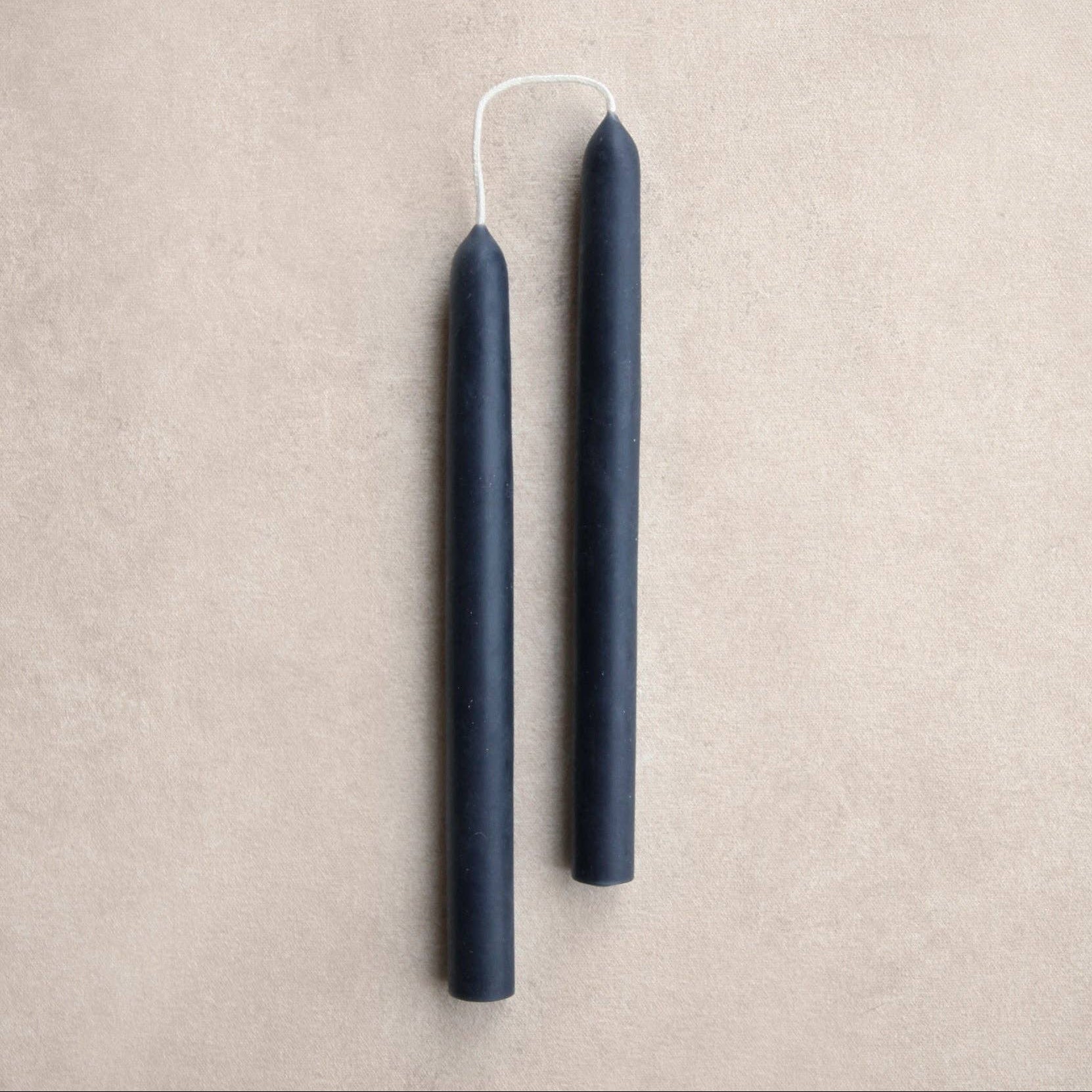 Two ink color taper candles with white wicks, standing vertically on a textured beige background.