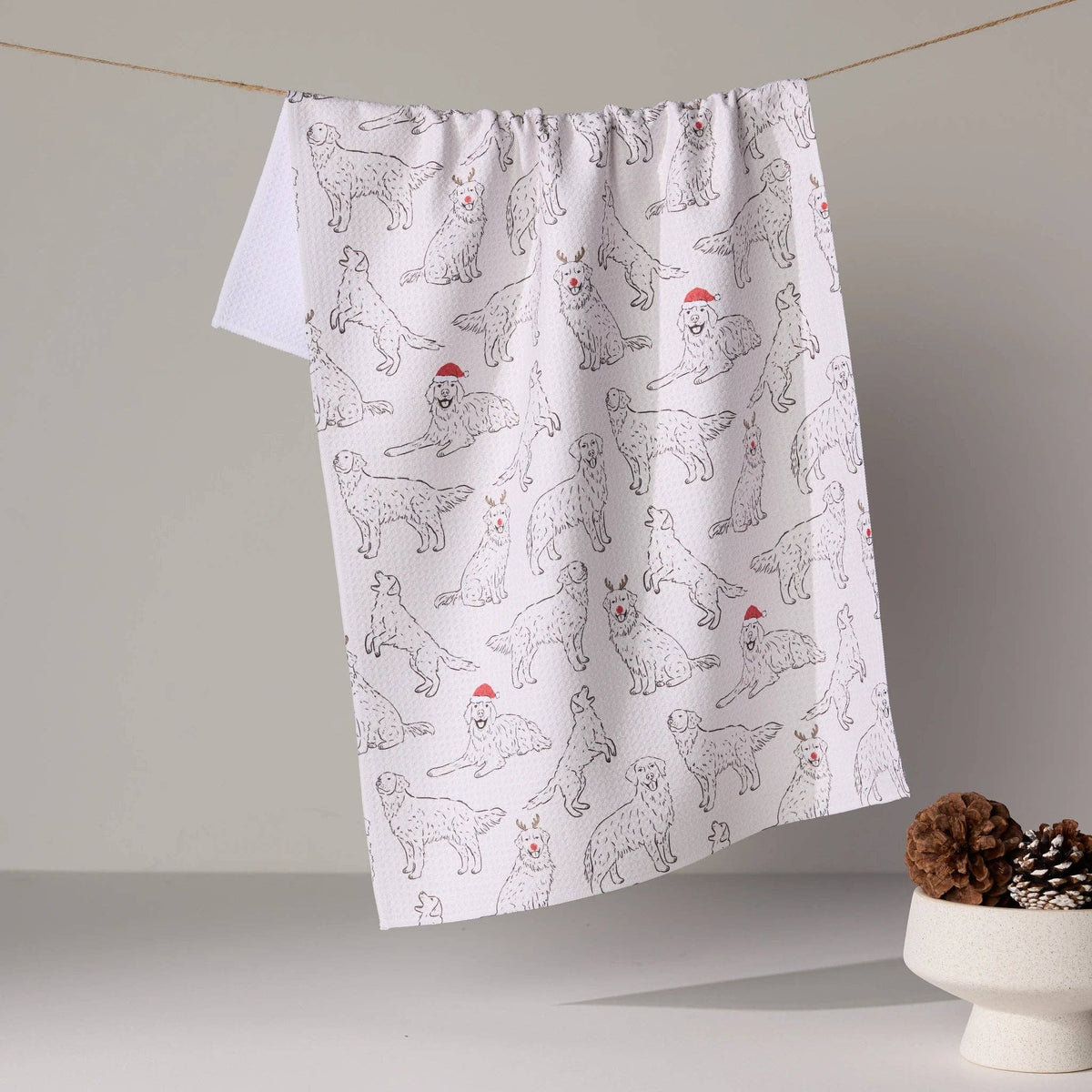 A white kitchen towel with a black line-art pattern of dogs wearing red Santa hats, hanging from a string in a minimalist studio setting.