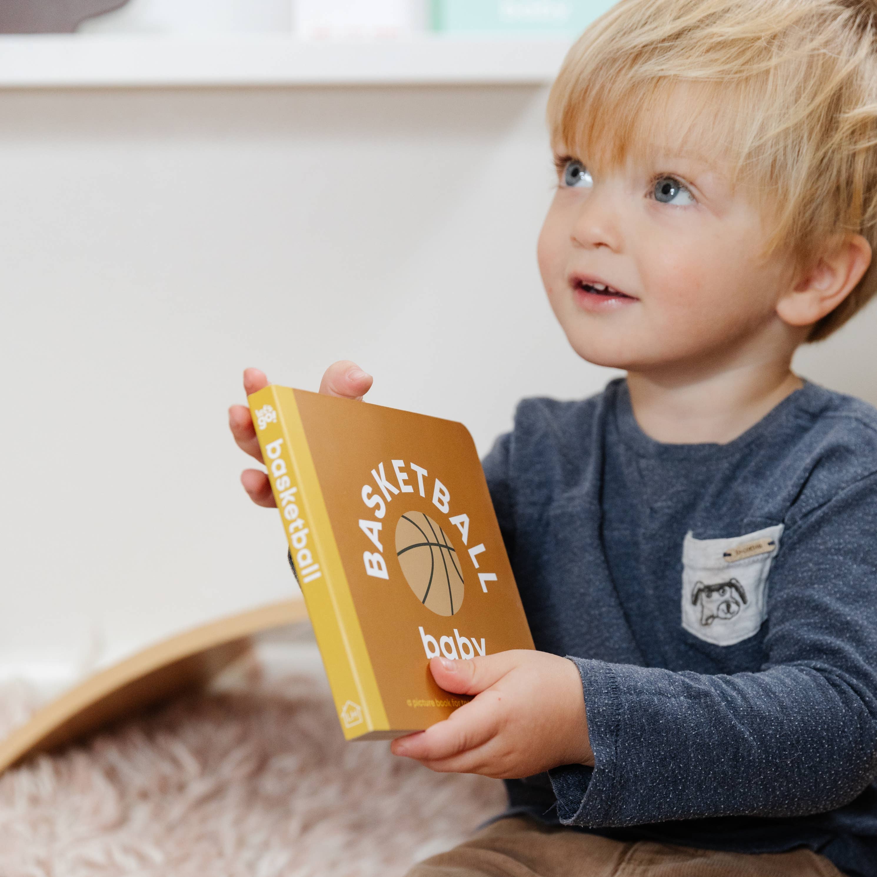 Child holding a book titled 'Basketball Baby' on a soft surface