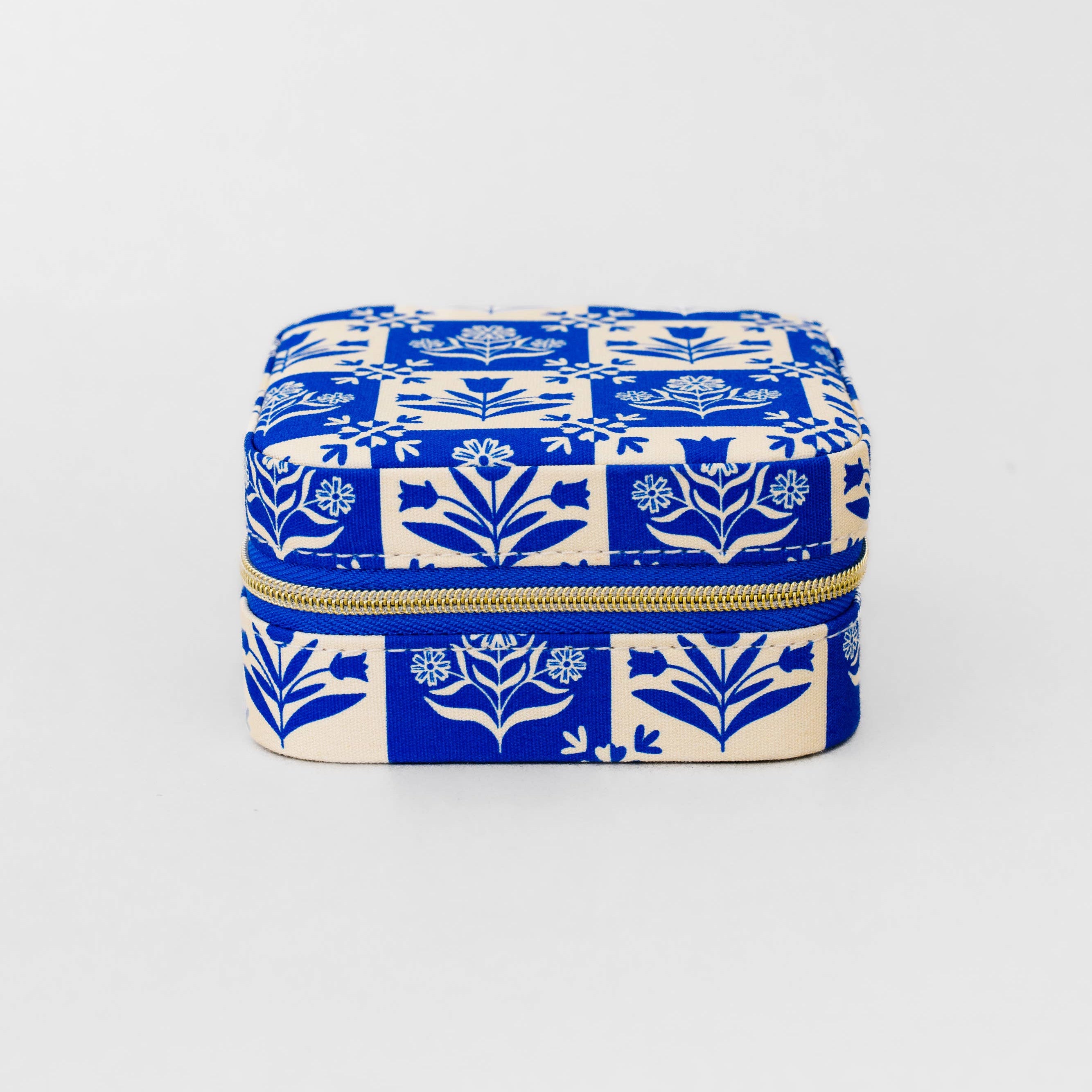 A square zippered case with a blue and cream floral block print pattern, featuring a gold zipper, set against a plain light gray background.