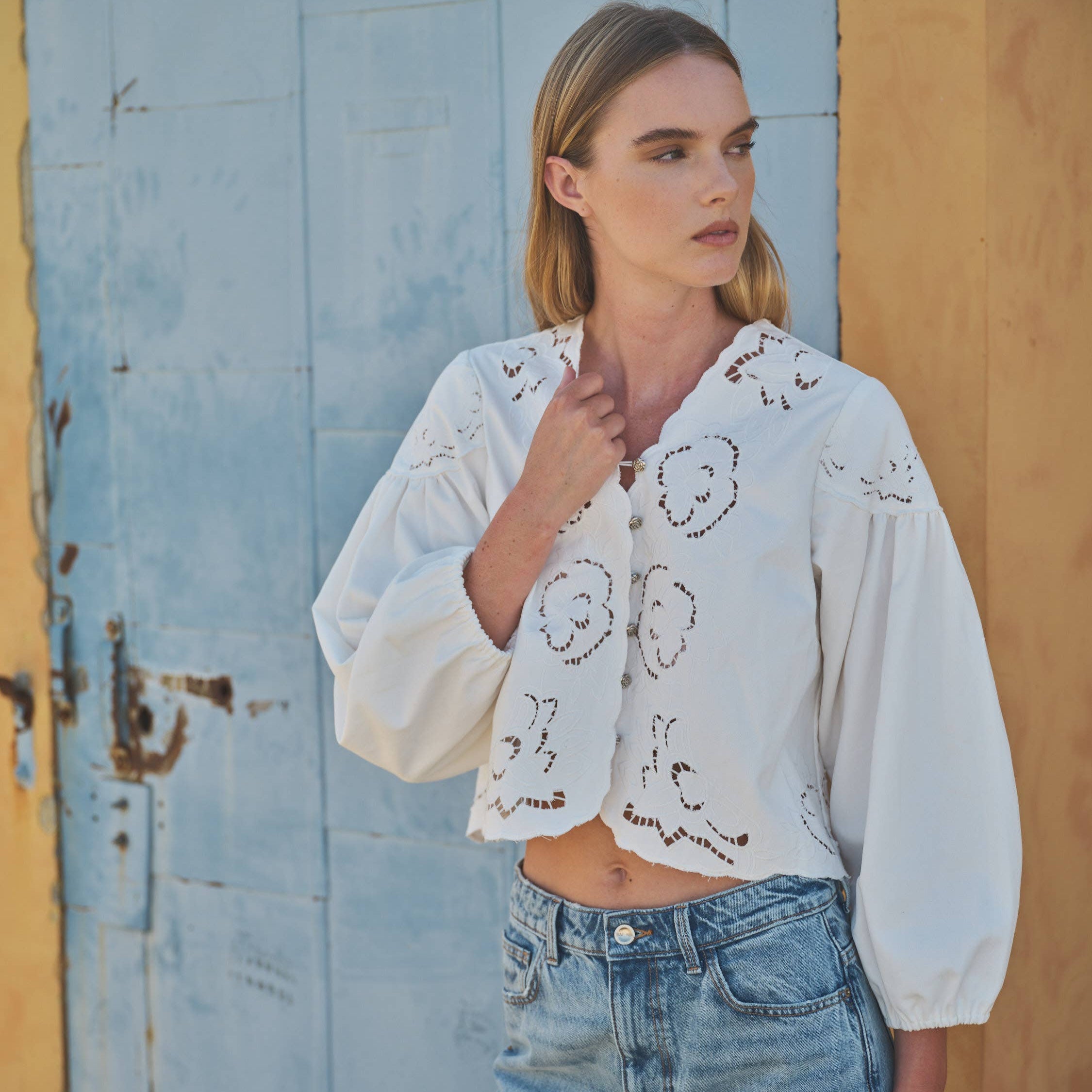Woman wearing a white embroidered blouse and blue jeans standing against a rustic wall.