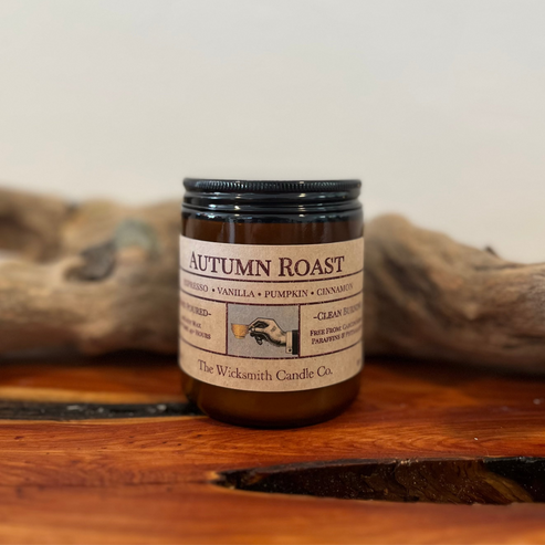 Amber glass candle jar labeled ‘Autumn Roast’ by The Wicksmith Candle Co., featuring scent notes of espresso, vanilla, pumpkin, and cinnamon, placed on a wooden surface with a piece of driftwood in the background