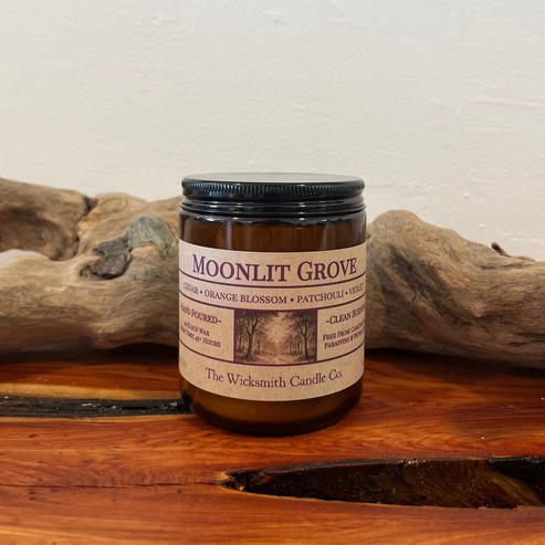 Amber glass candle jar labeled ‘Moonlit Grove’ by The Wicksmith Candle Co., featuring scent notes of cedar, orange blossom, patchouli, and violet placed on a wooden surface with a piece of driftwood in the background