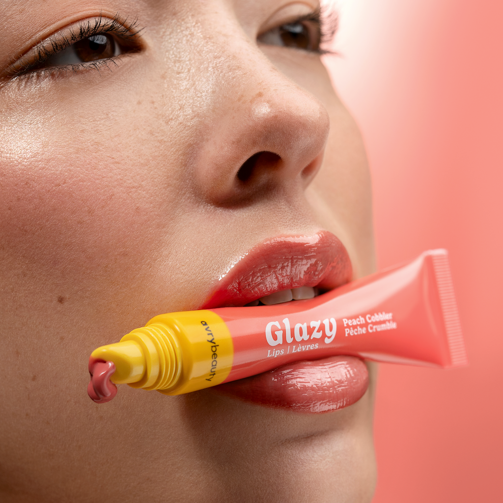 Person holding a tube of Glazy lip balm close to their mouth against a pink background