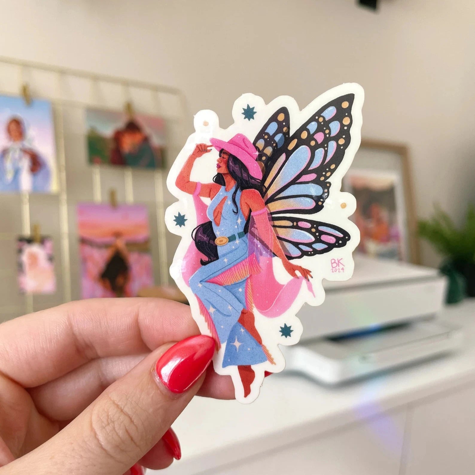 This sticker has a glossy vinyl finish and a clear back once taken off the backing paper. A black cowgirl with long black hair wearing a pink cowgirl hat and a denim jumpsuit with fringe along the legs. She has large butterfly wings attached to her back.