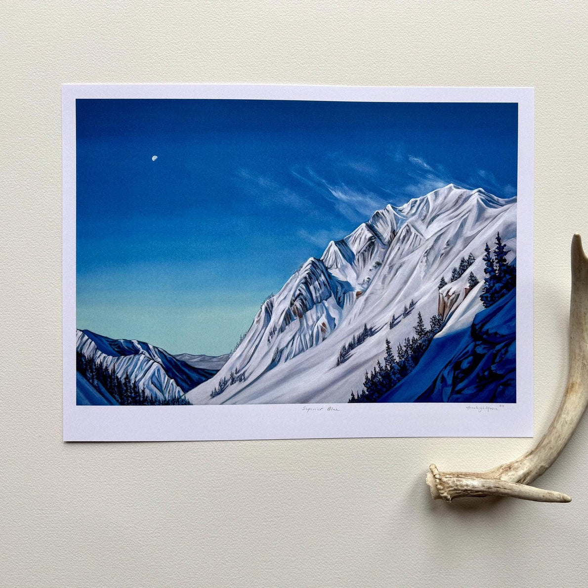Print against a white wall with a decorative antler. Painting is done is vibrant rich blues, with a dramatic mountain range, rich shadows, snow whips, and a stark tiny moon in the sky.