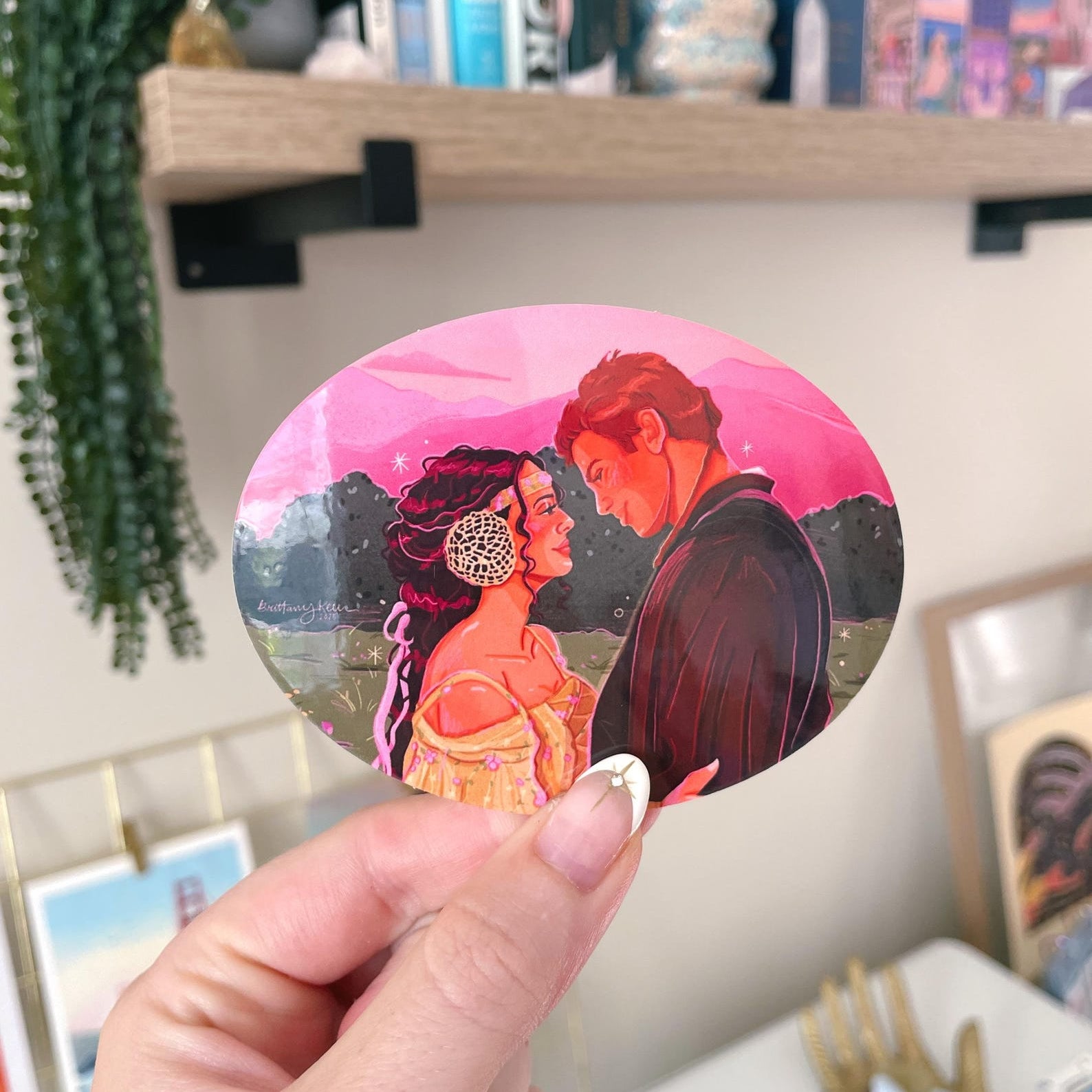 a hand holding a sticker showing a drawing of a man and woman embracing and looking at each other lovingly, with a pink sky and green scenery behind them