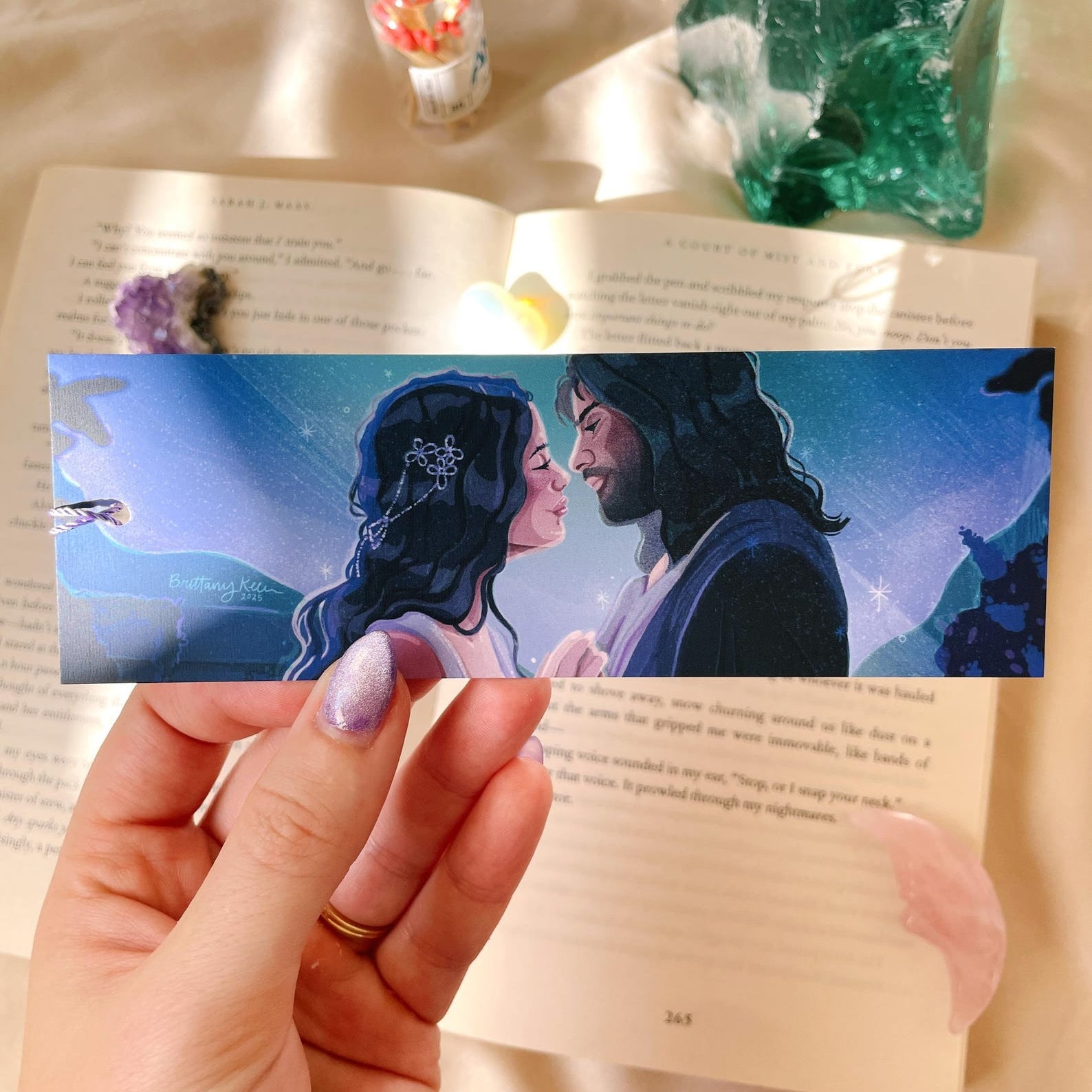 a hand over a book holding a rectangular bookmark featuring a man and woman holding hands looking at each other lovingly with a blue and purple starry scene behind them