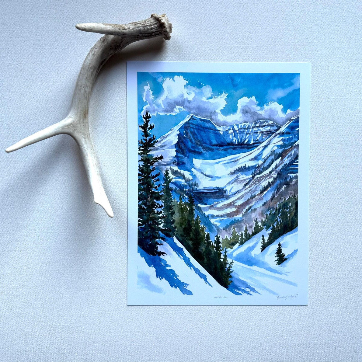 Dramatic painting in blues with green pines of Mount Timpanogos. Print is against a white wall with a decorative antler.