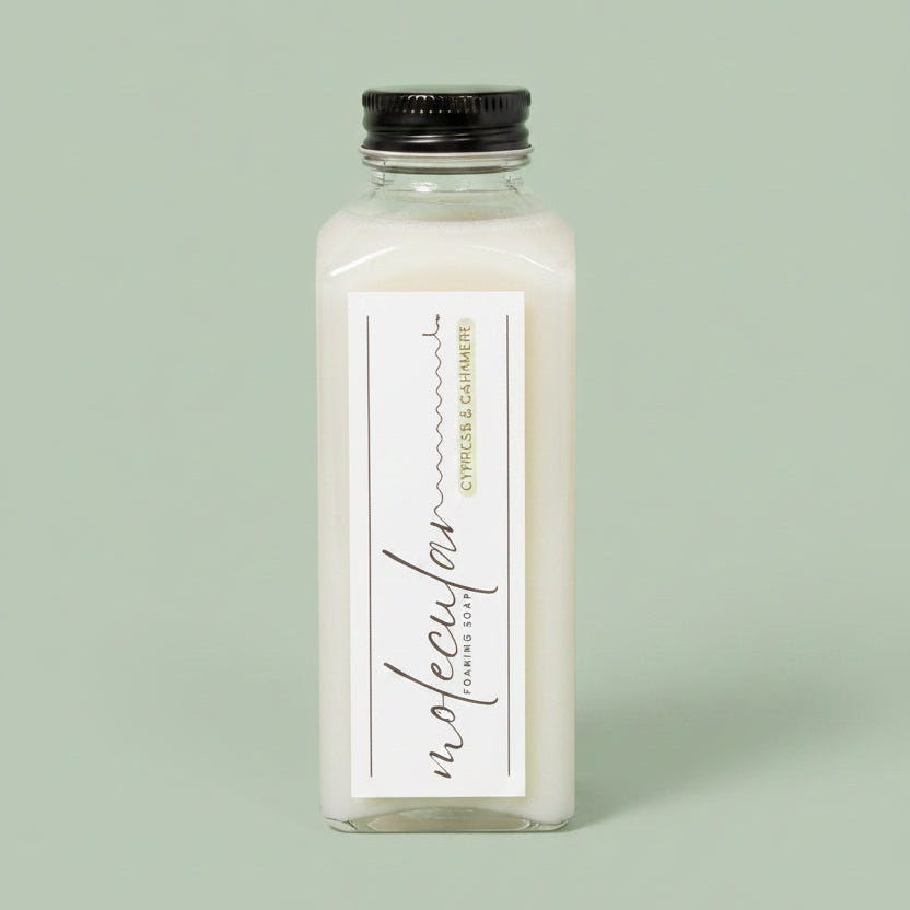 A hand holds a clear rectangular bottle of Molecular Foaming Soap labeled Cypress and Cashmere. The bottle contains a pale liquid and is photographed against a soft green background