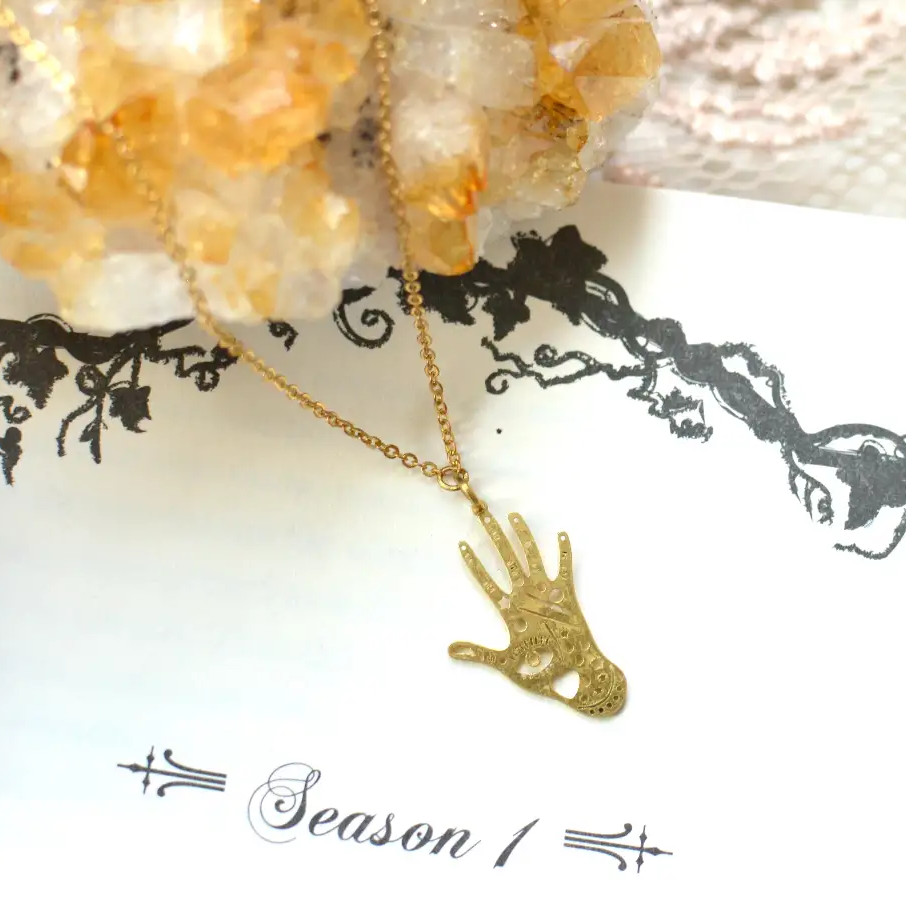 A gold-tone hand-shaped pendant necklace featuring intricate engraved patterns and a small heart cutout in the palm. The delicate gold chain is displayed on a white jewelry card with vintage-inspired black floral detailing, accented by a golden crystal cluster in the background.