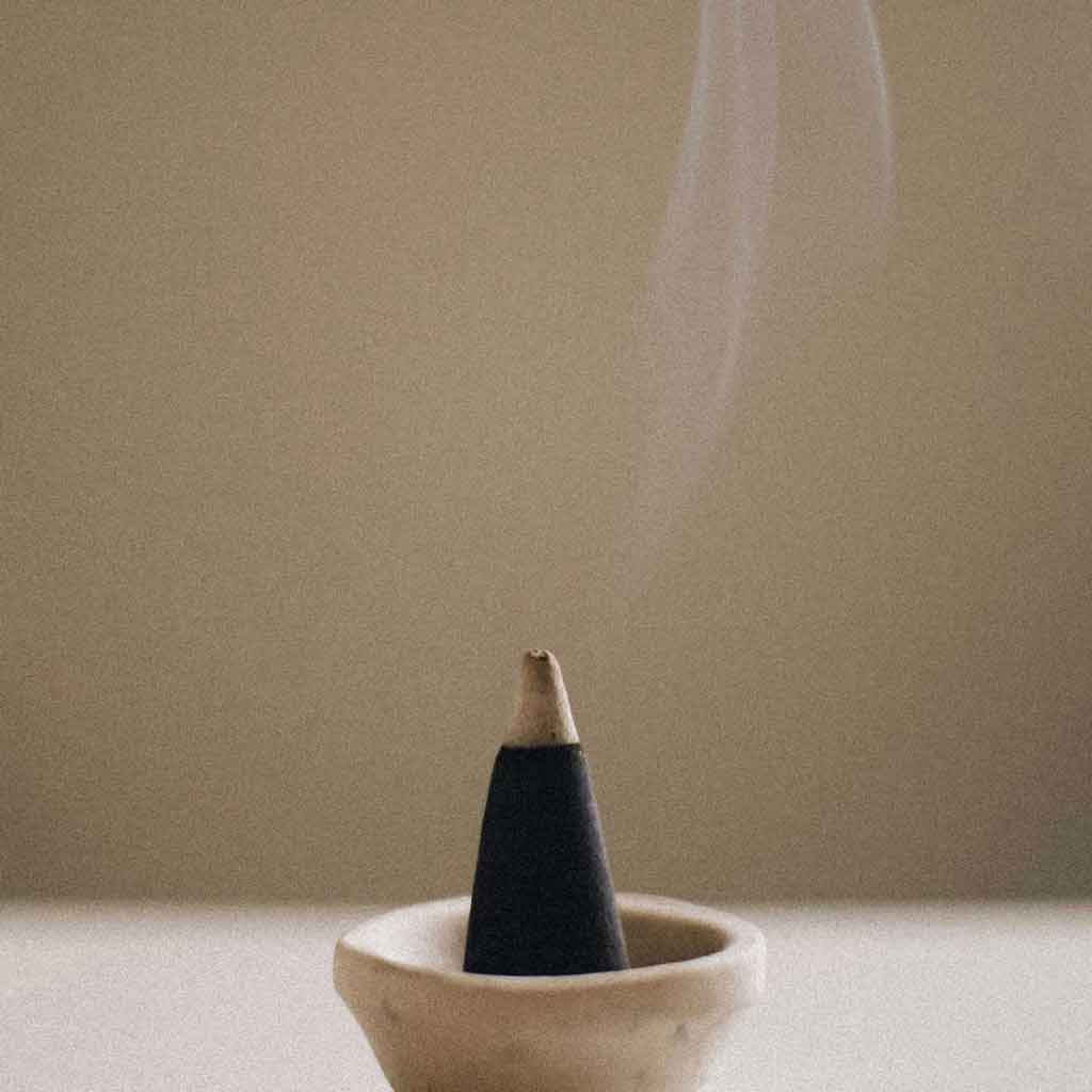 A close-up photograph shows a single black charcoal incense cone with a light beige tip, burning and emitting a thin stream of white smoke, placed upright in a small, shallow, off-white ceramic holder with a textured, natural stone-like appearance. The soft neutral background emphasizes the gentle curl of smoke rising from the cone in a calm, minimalist composition.