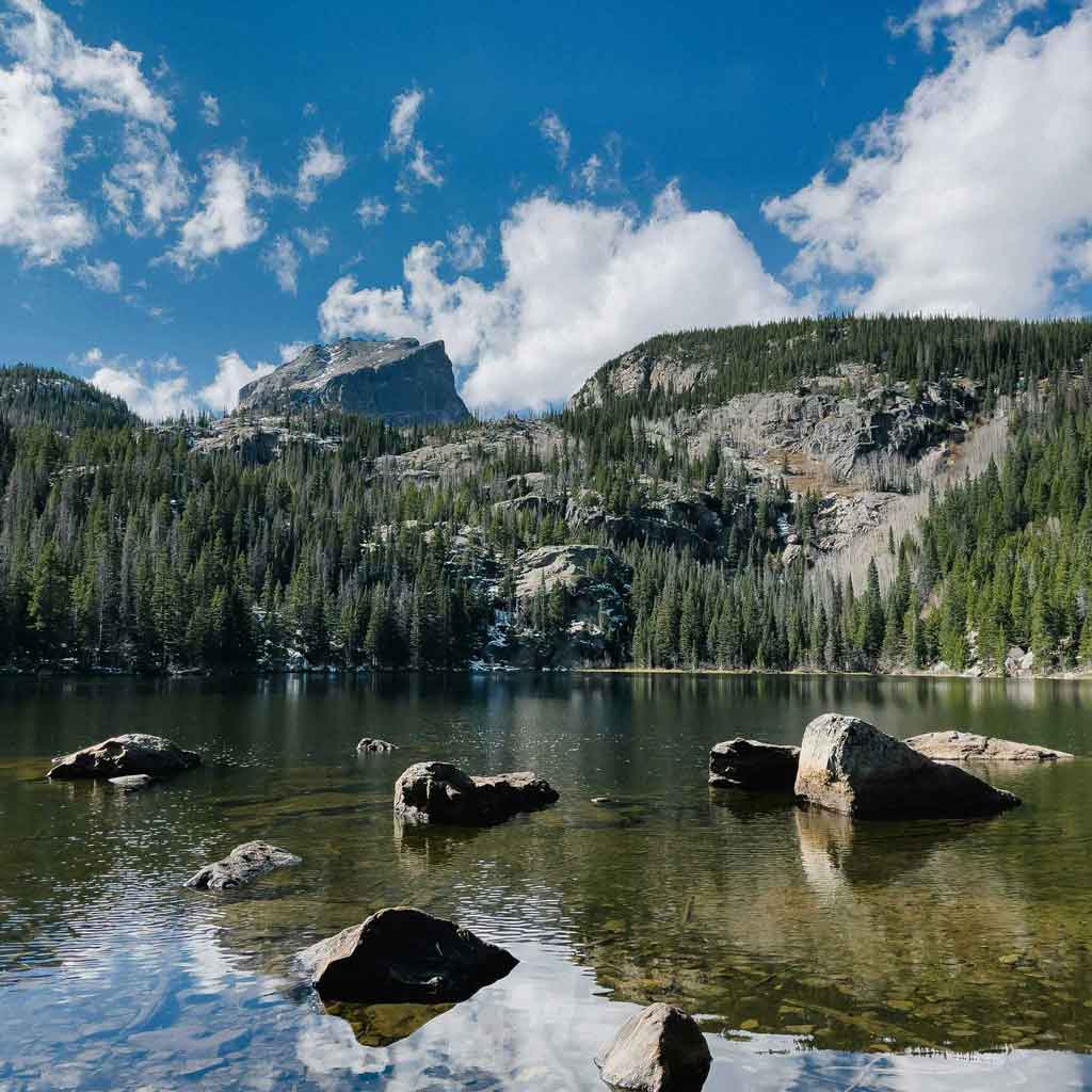 A serene alpine lake in Utah reflects a dramatic rocky mountain peak under a bright blue sky scattered with white clouds. The foreground features clear green-tinted water with several large granite boulders emerging from the surface, while dense evergreen forests cover the surrounding slopes and ridges.