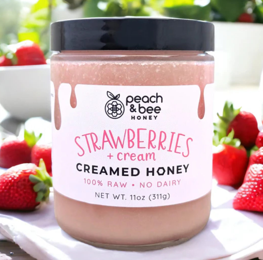 A jar of Peach & Bee creamed honey with strawberries and cream flavor, placed on a table with whole strawberries and a bowl in the background.