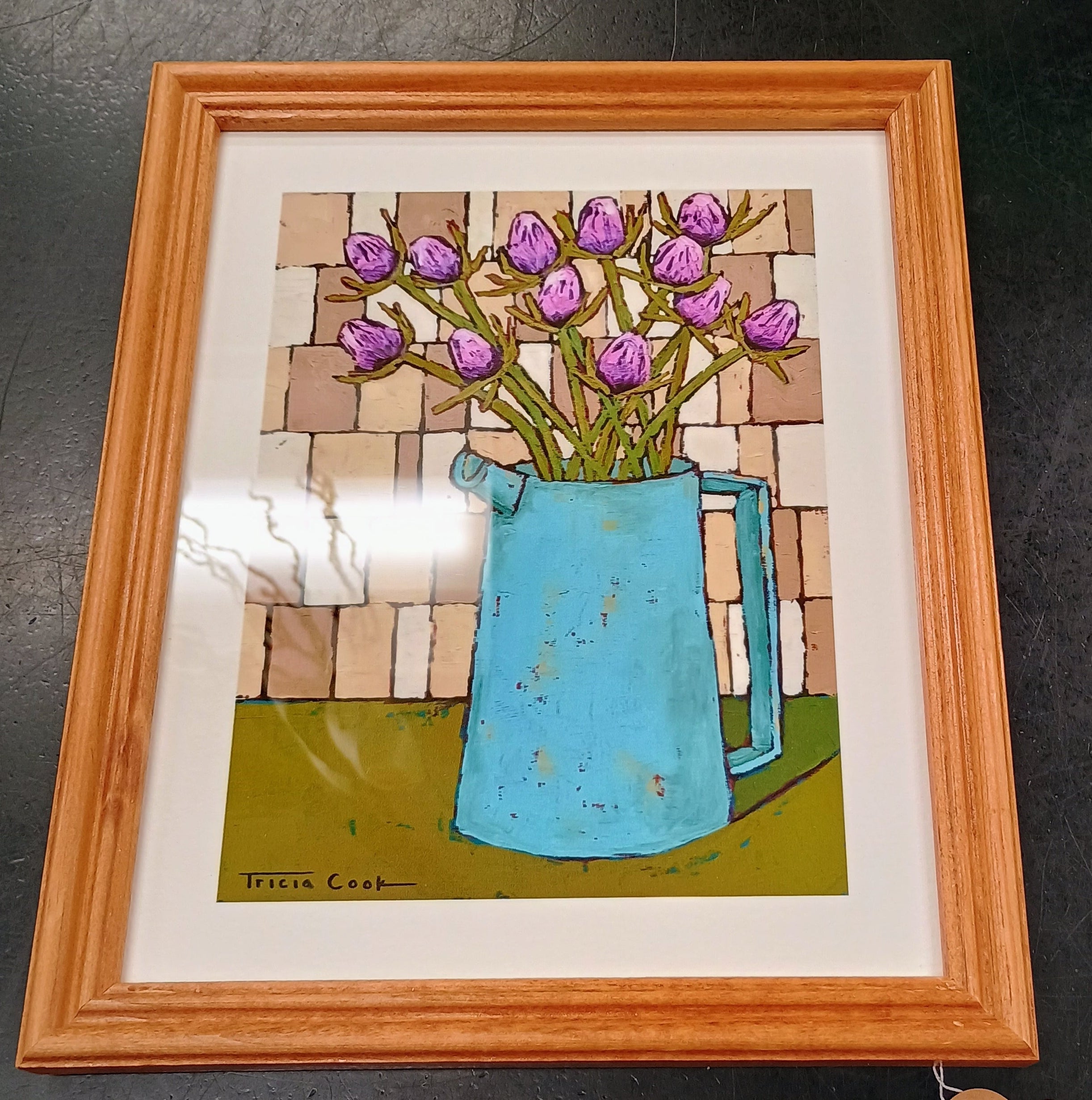 Framed print of purple thistle buds in a sky blue pitcher, on a green table top, against a light brown tile wall. Frame is oak