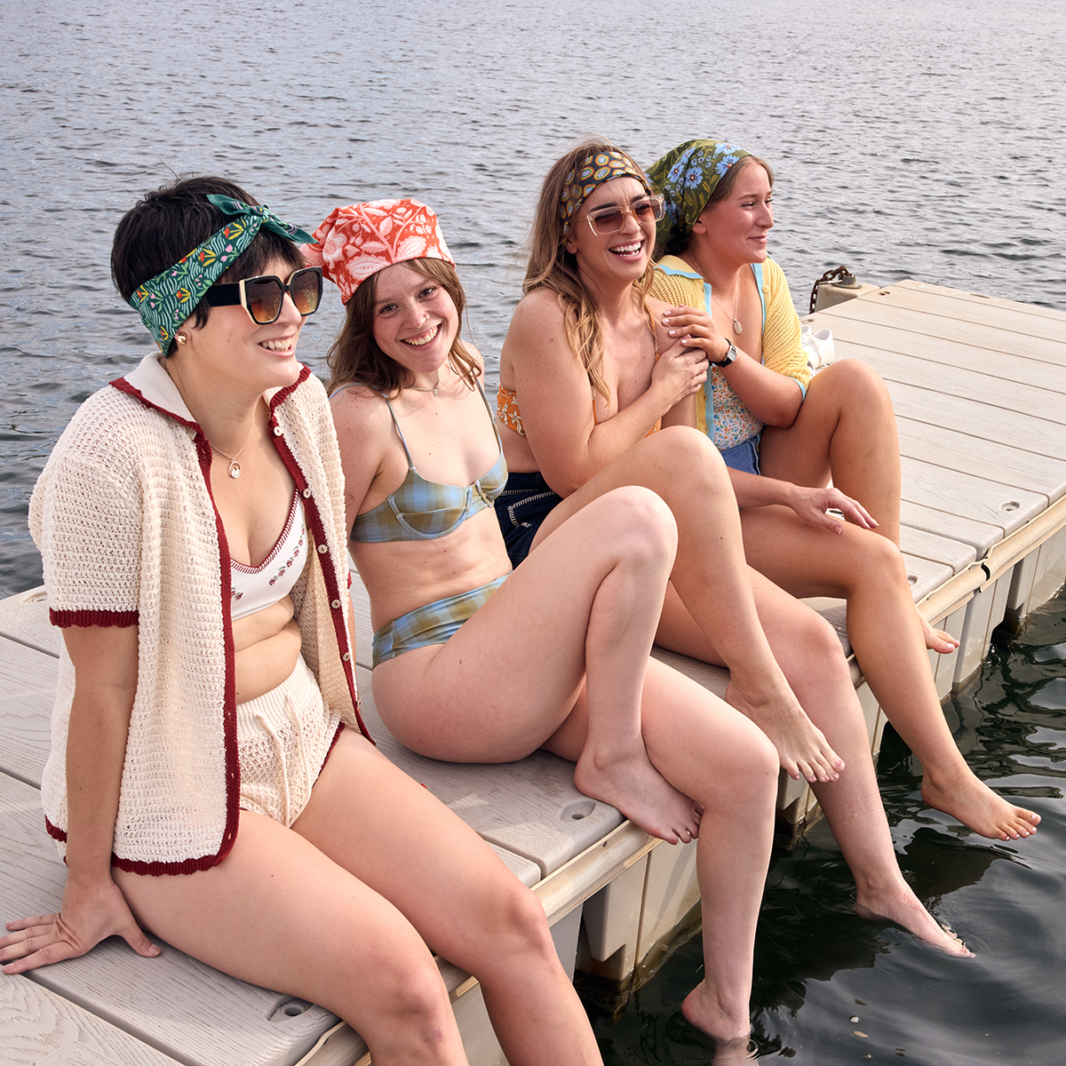 Four women sitting on a dock by a lake, wearing swimsuits and sunglasses.