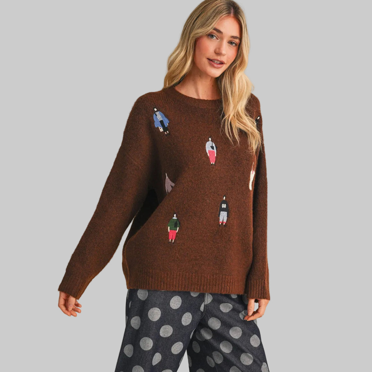 A woman wearing a broen knit sweater with small colorful women dressed in fun clothing patches, paired with gray polka-dot pants, against a plain beige studio background.